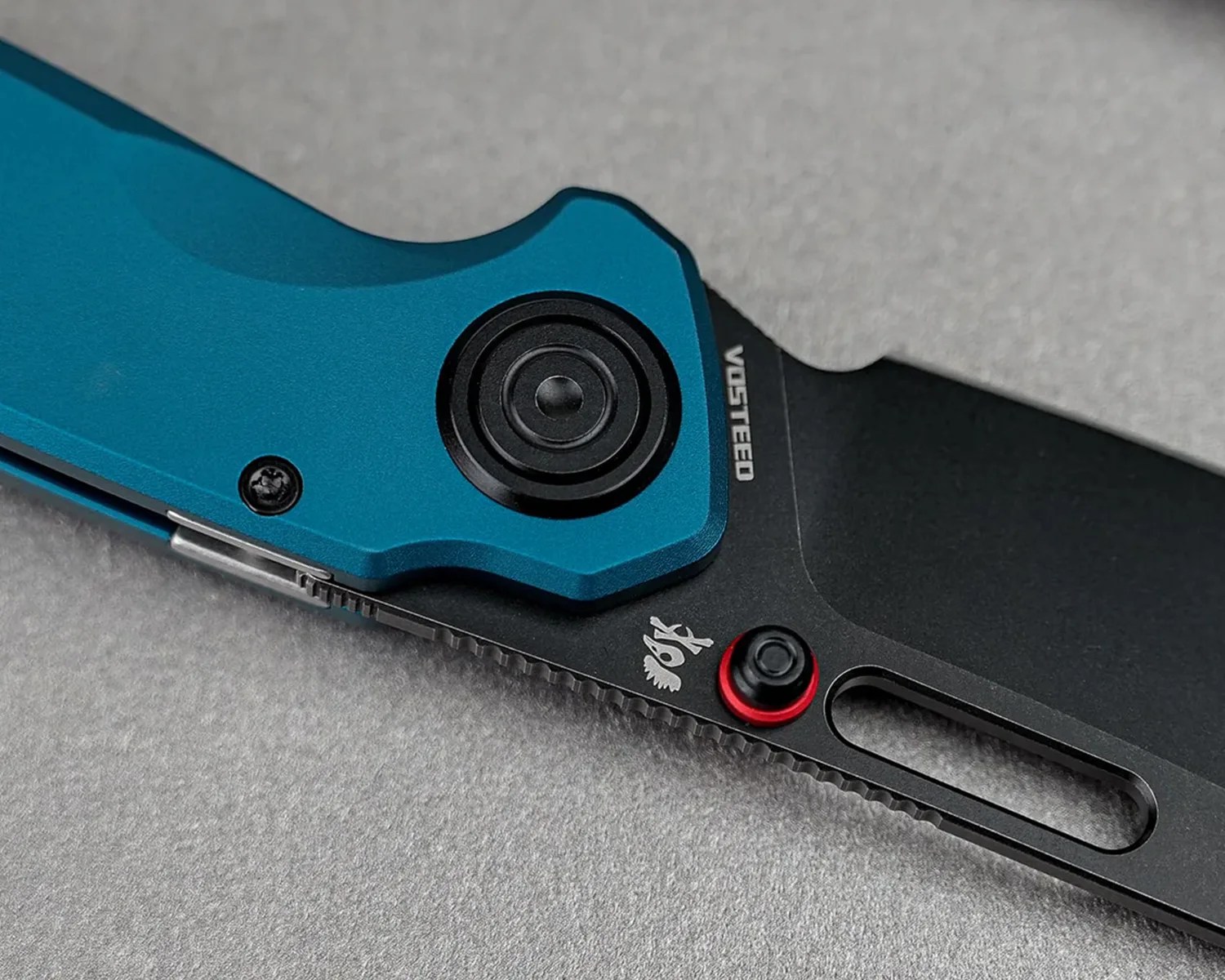 Close-up of a black and blue folding knife handle with a thumb stud and the brand name "Vosteed" visible.