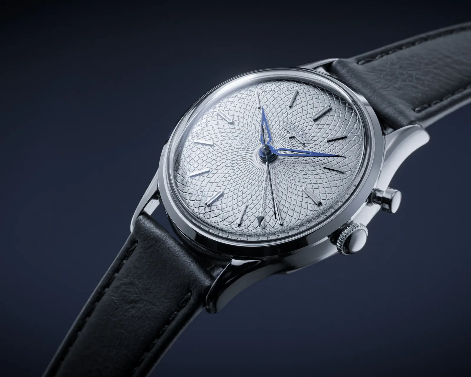 Silver wristwatch with intricate geometric patterned dial, blue hands, and black leather strap on dark background.