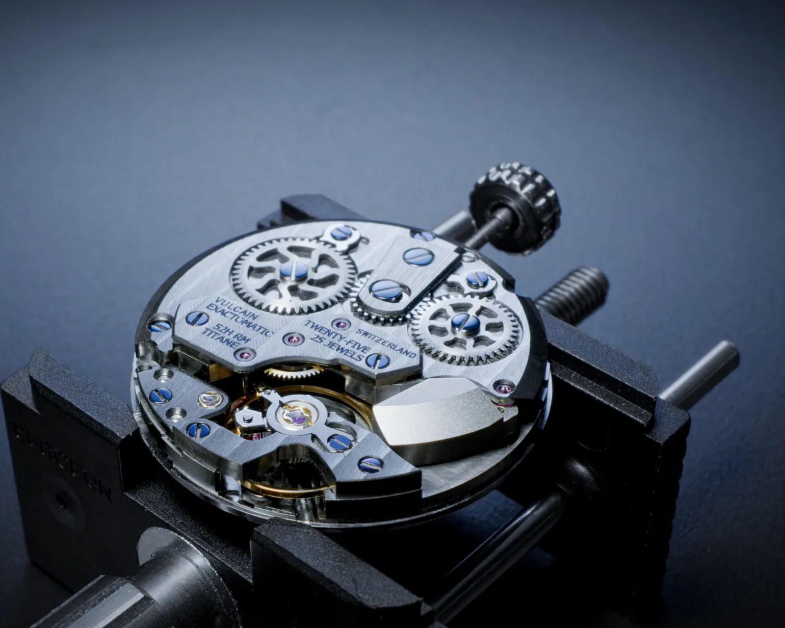 Close-up of a mechanical watch movement with visible gears, screws, and inscriptions held in a black watchmaker's holder.