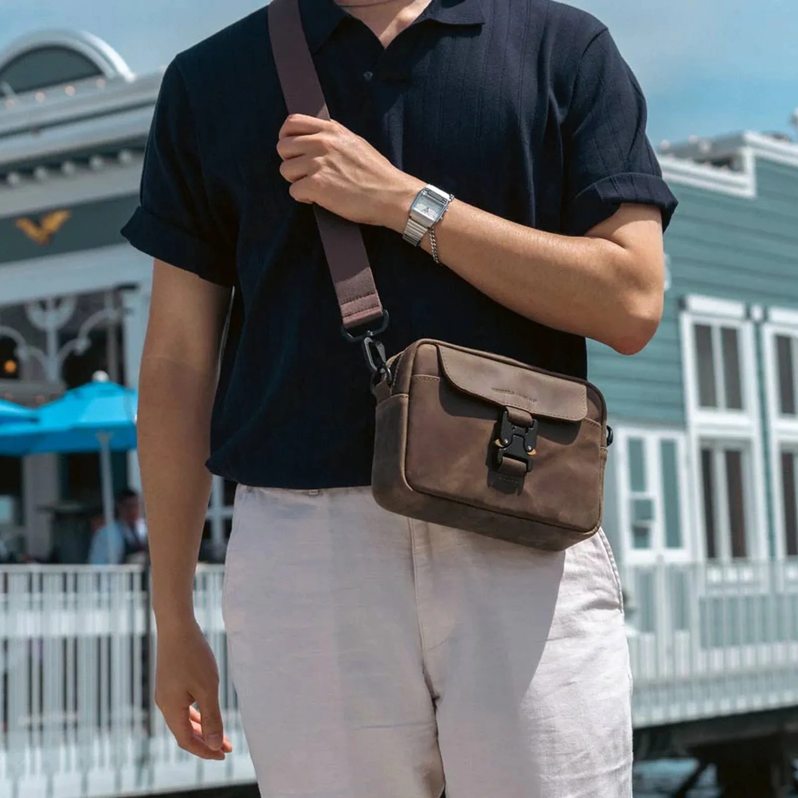 Brown leather crossbody bag with black buckle worn over a navy short-sleeve shirt and light pants.
