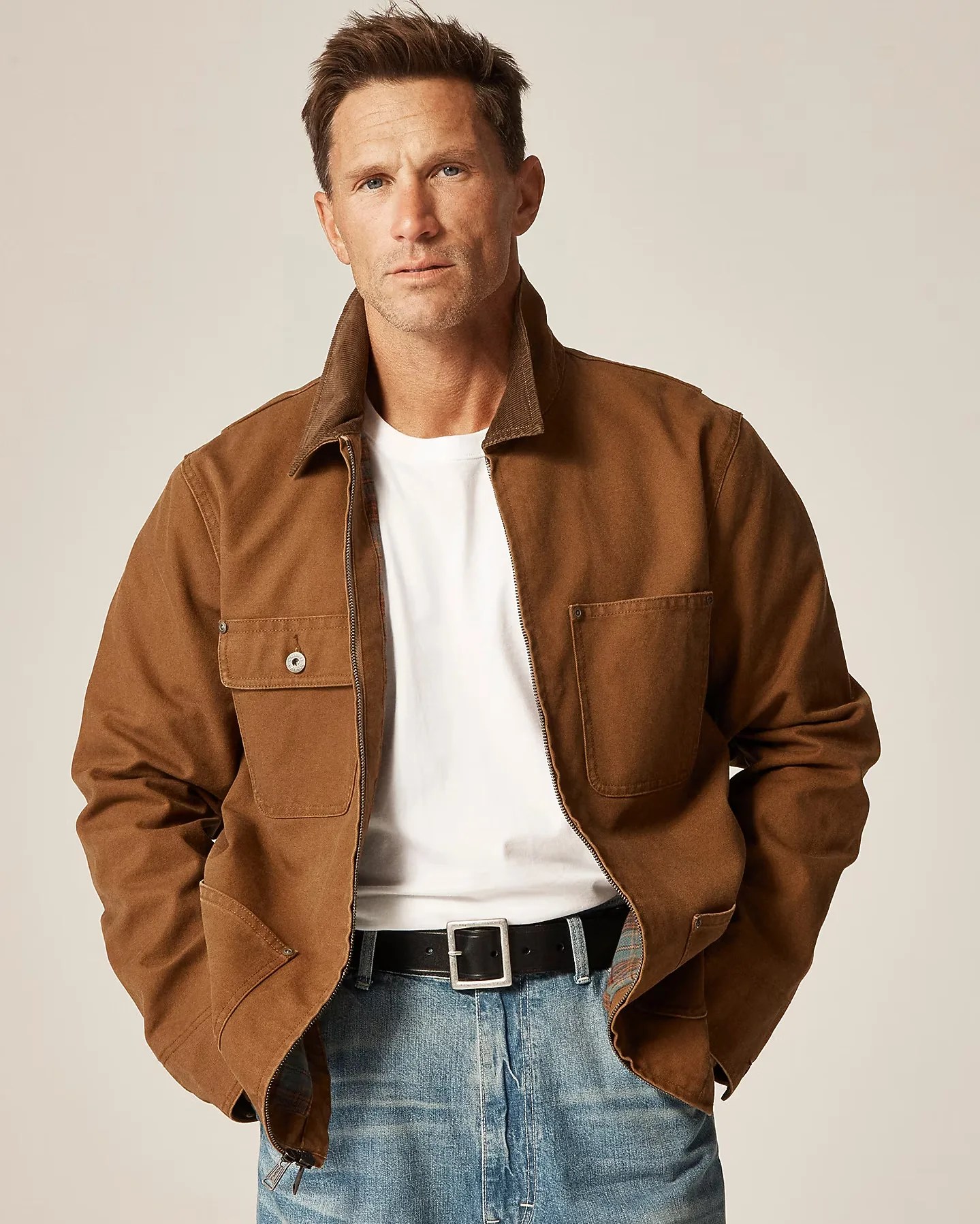 Man wearing a brown zip-up jacket with flap and patch pockets over a white t-shirt and blue jeans.