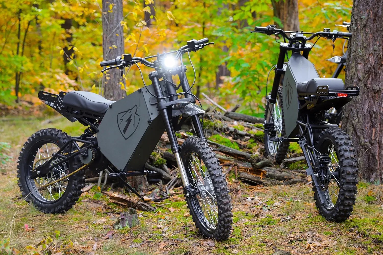 Two black electric dirt bikes with knobby tires and angular battery compartments parked on forest ground with autumn foliage.