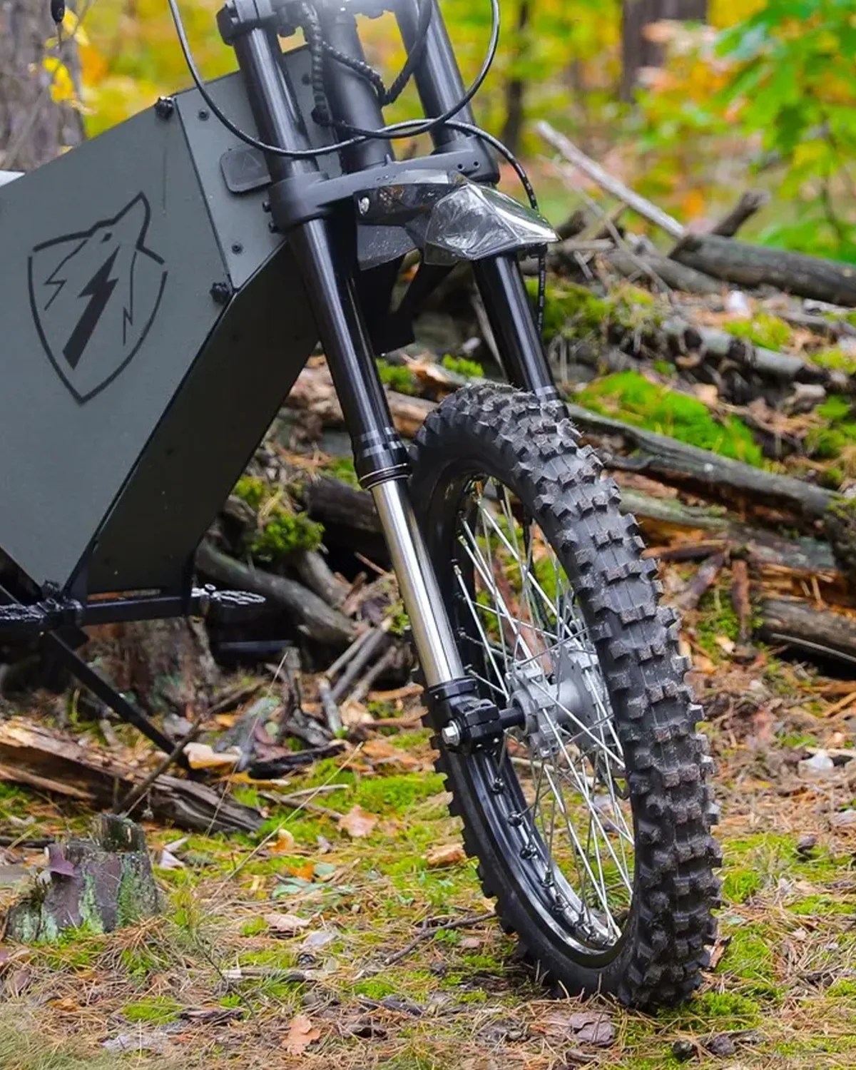 Front wheel and suspension fork of a black electric dirt bike with knobby tire on a forest floor.