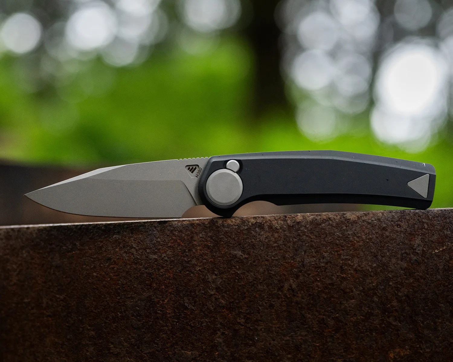 Folding knife with a gray blade marked M390 and a black handle resting on a rusty metal surface.
