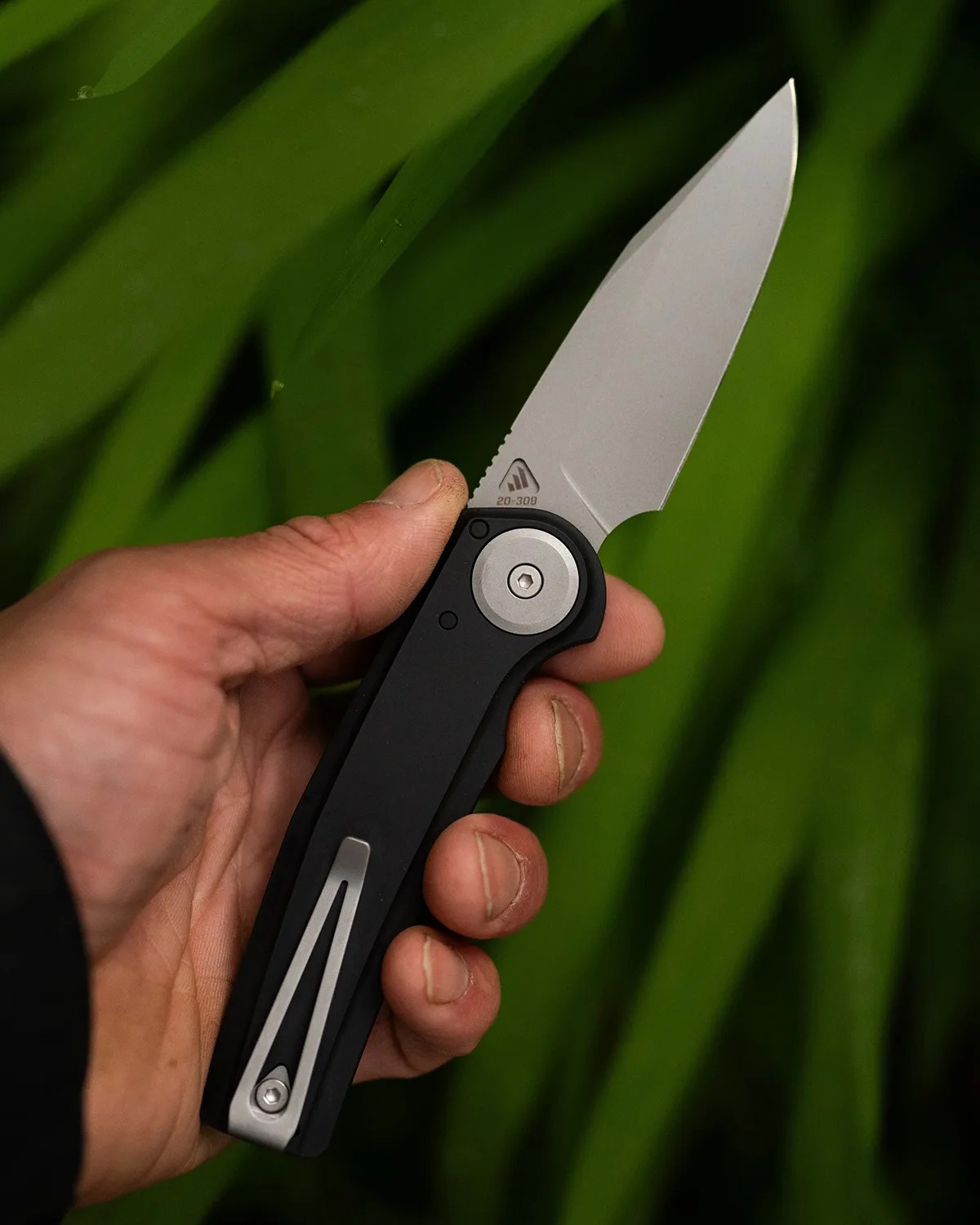 Hand holding a folding knife with a black handle and silver blade against a green leafy background.