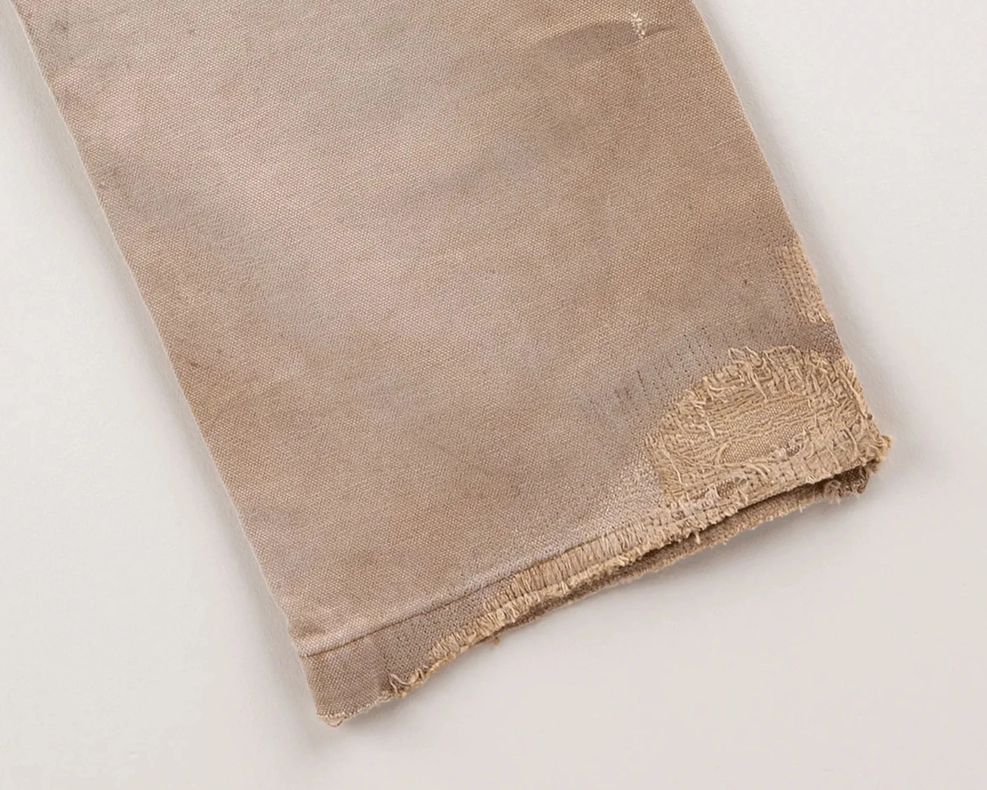 Beige fabric with frayed edges and a worn patch on a light surface.