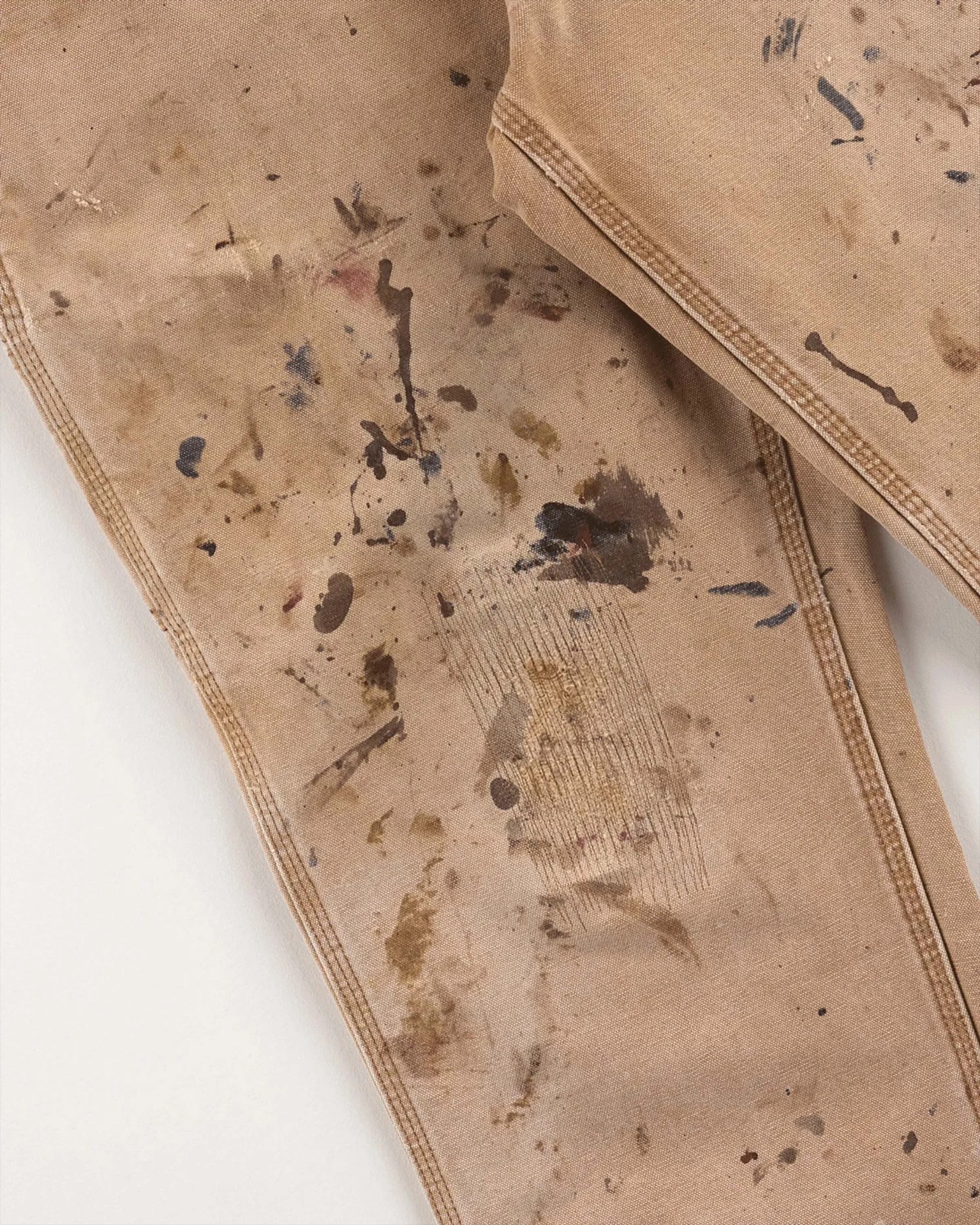 Tan canvas pants with multiple paint stains and visible stitching on a white background.