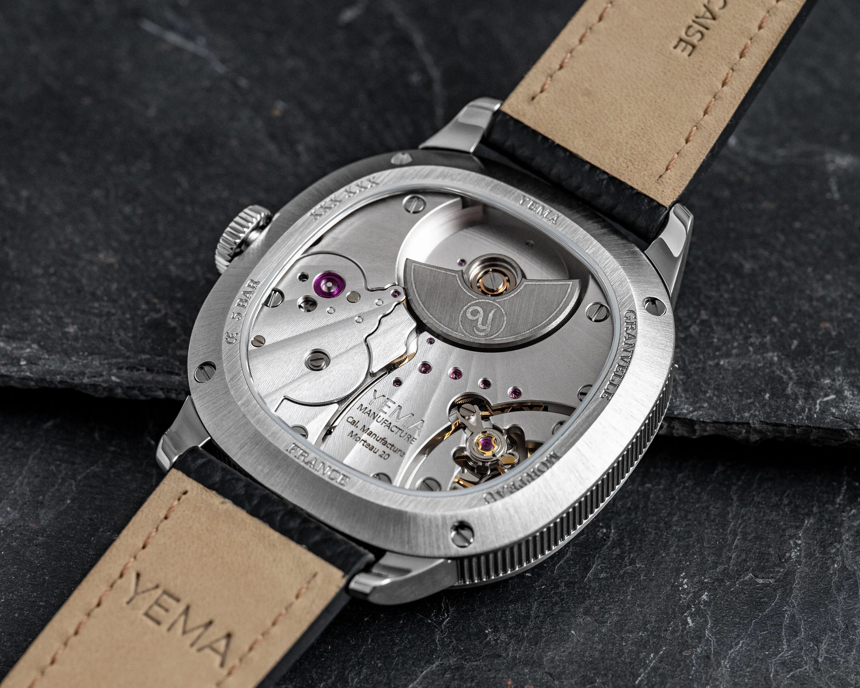 Close-up of the back of a Yema wristwatch showing its mechanical movement and beige leather strap on a dark stone surface.