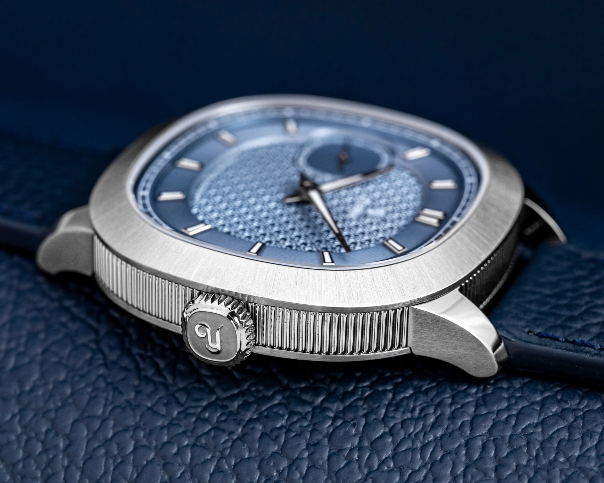 Silver wristwatch with textured blue dial and ridged crown on a dark blue leather surface.