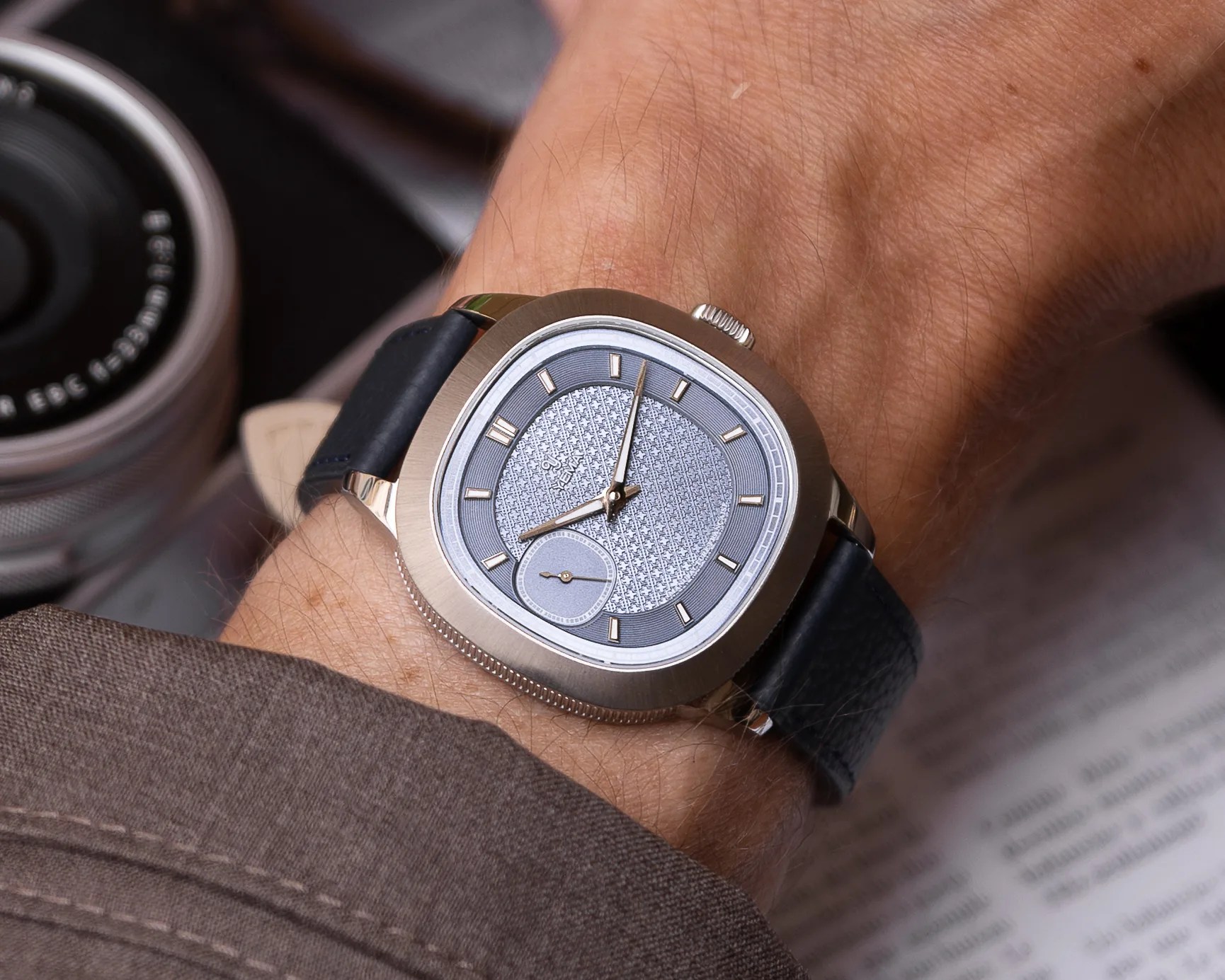 Square silver wristwatch with textured gray dial and black leather strap worn on a wrist.