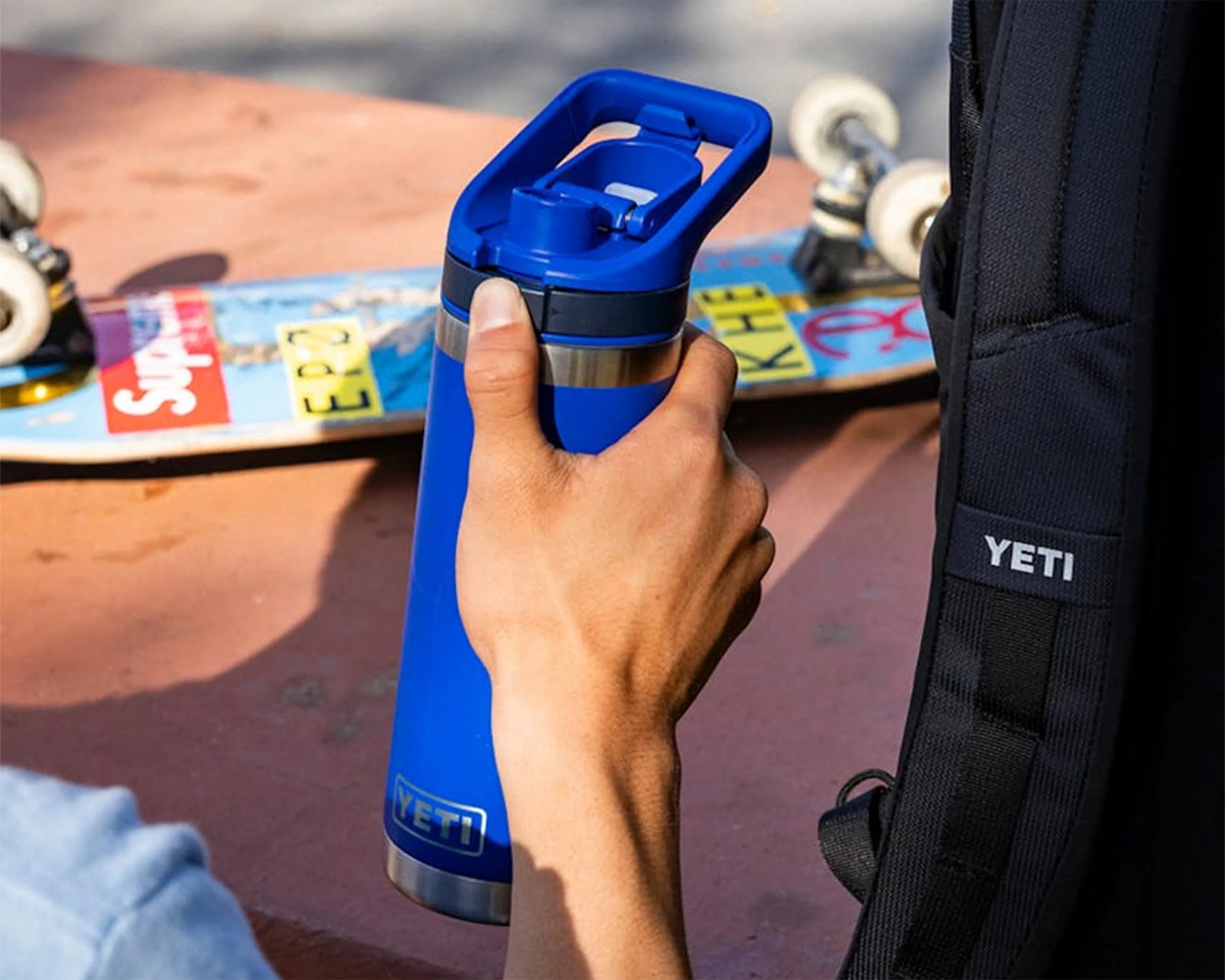 Hand holding a blue YETI insulated water bottle with a skateboard and black YETI bag in the background.