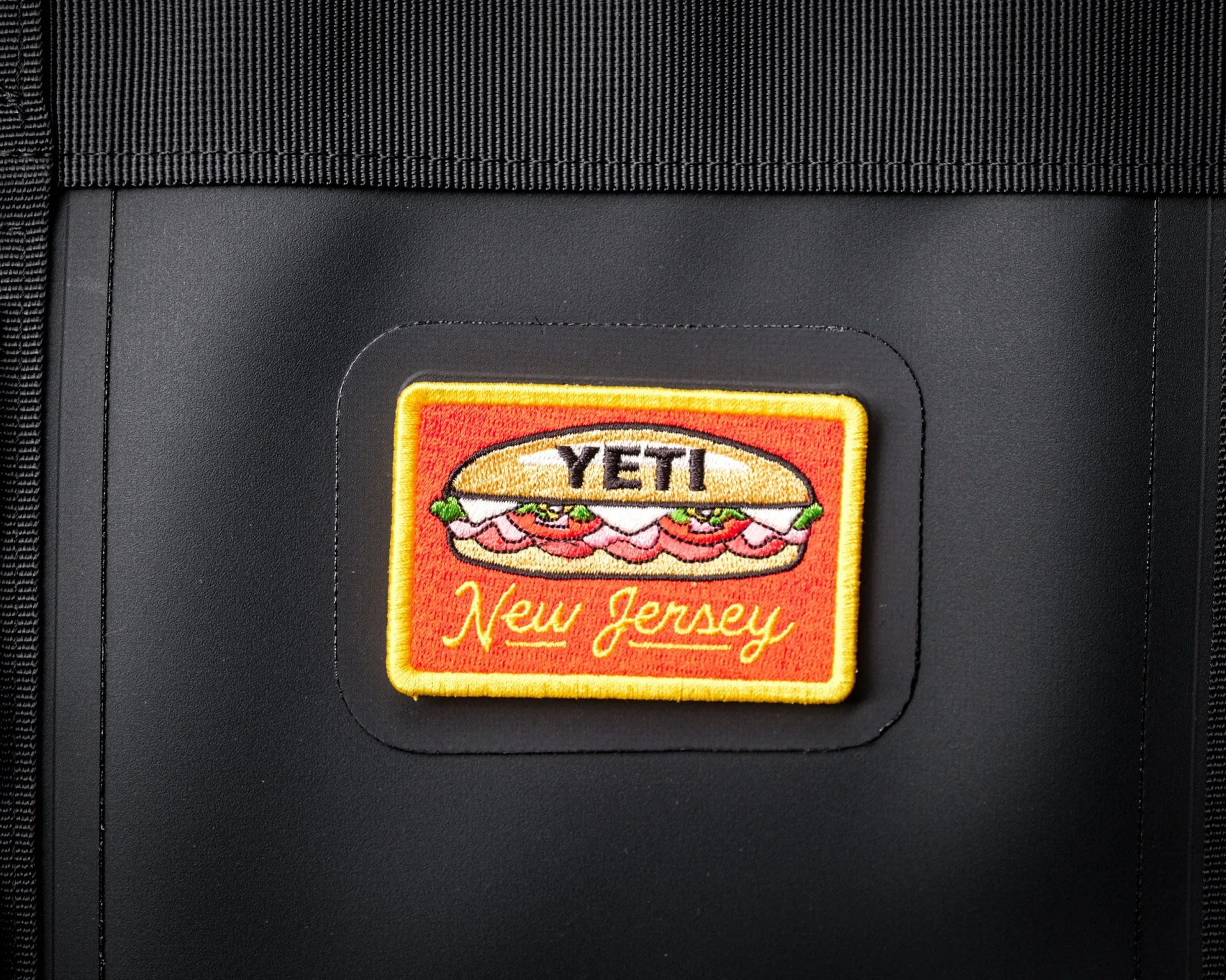 Black fabric and leather surface with a rectangular embroidered patch showing a sandwich and the text "YETI New Jersey.