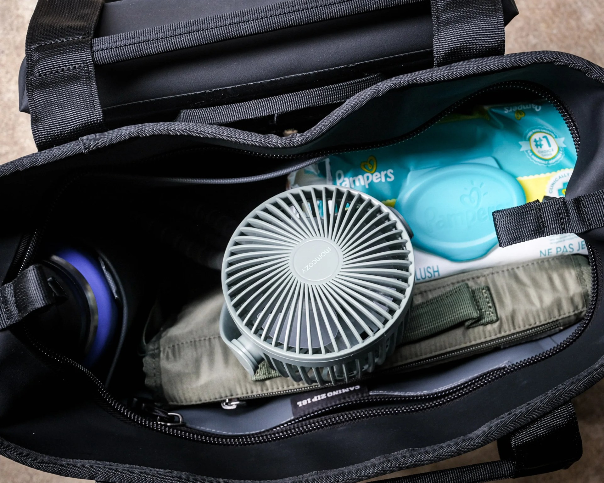 Black bag with a gray portable fan, Pampers wipes, and a folded khaki fabric item inside.