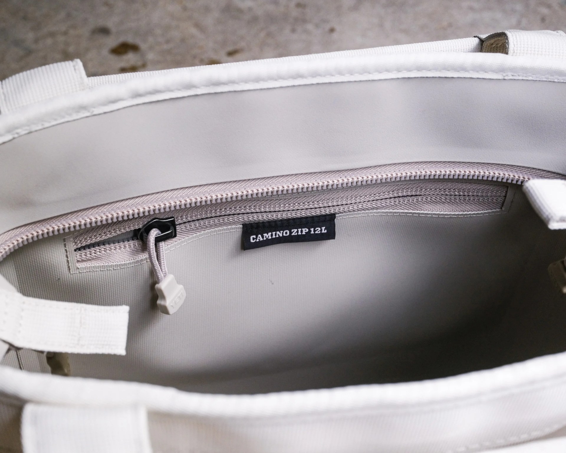 Interior of a light-colored bag with a zippered pocket labeled "CAMINO ZIP 12L" and a white zipper pull.