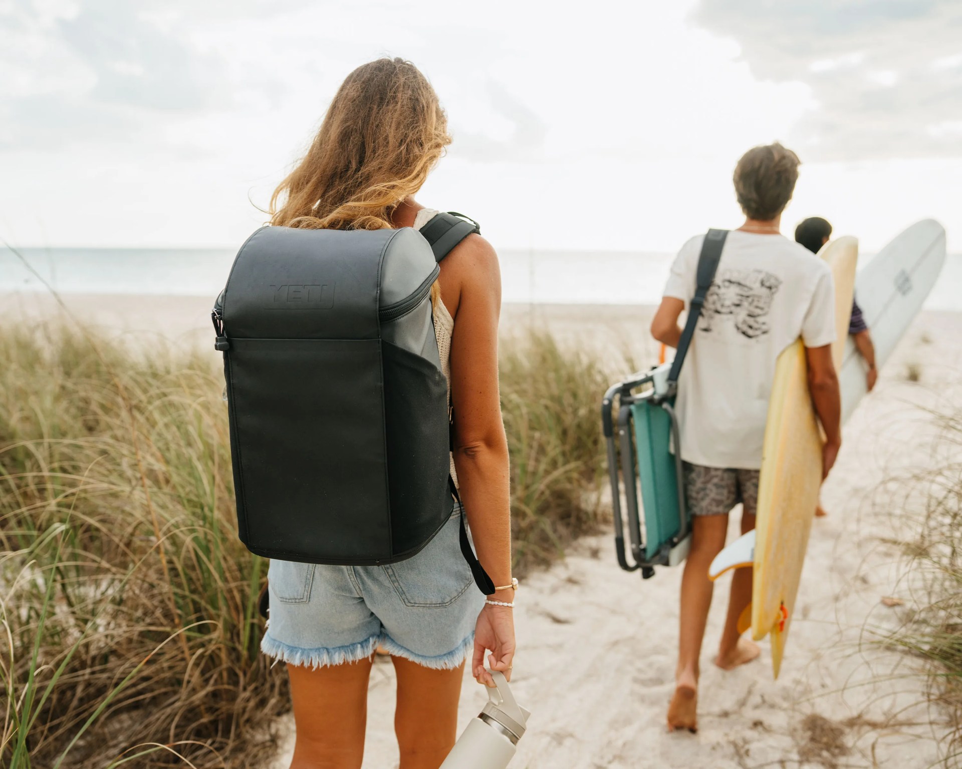 Person wearing a black YETI backpack and denim shorts walking on a sandy beach path toward the ocean.