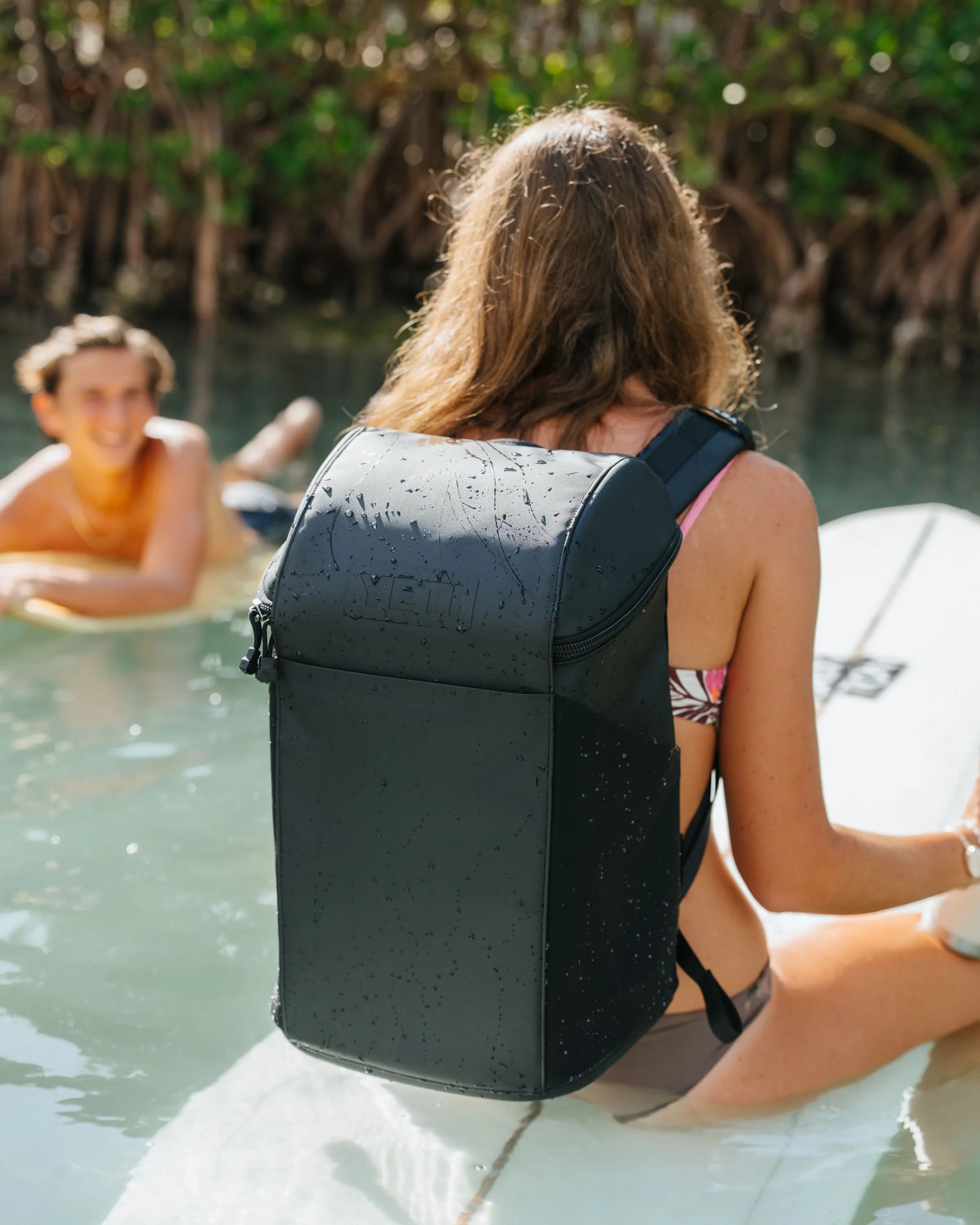 Black waterproof YETI backpack worn by a person sitting on a paddleboard in water.