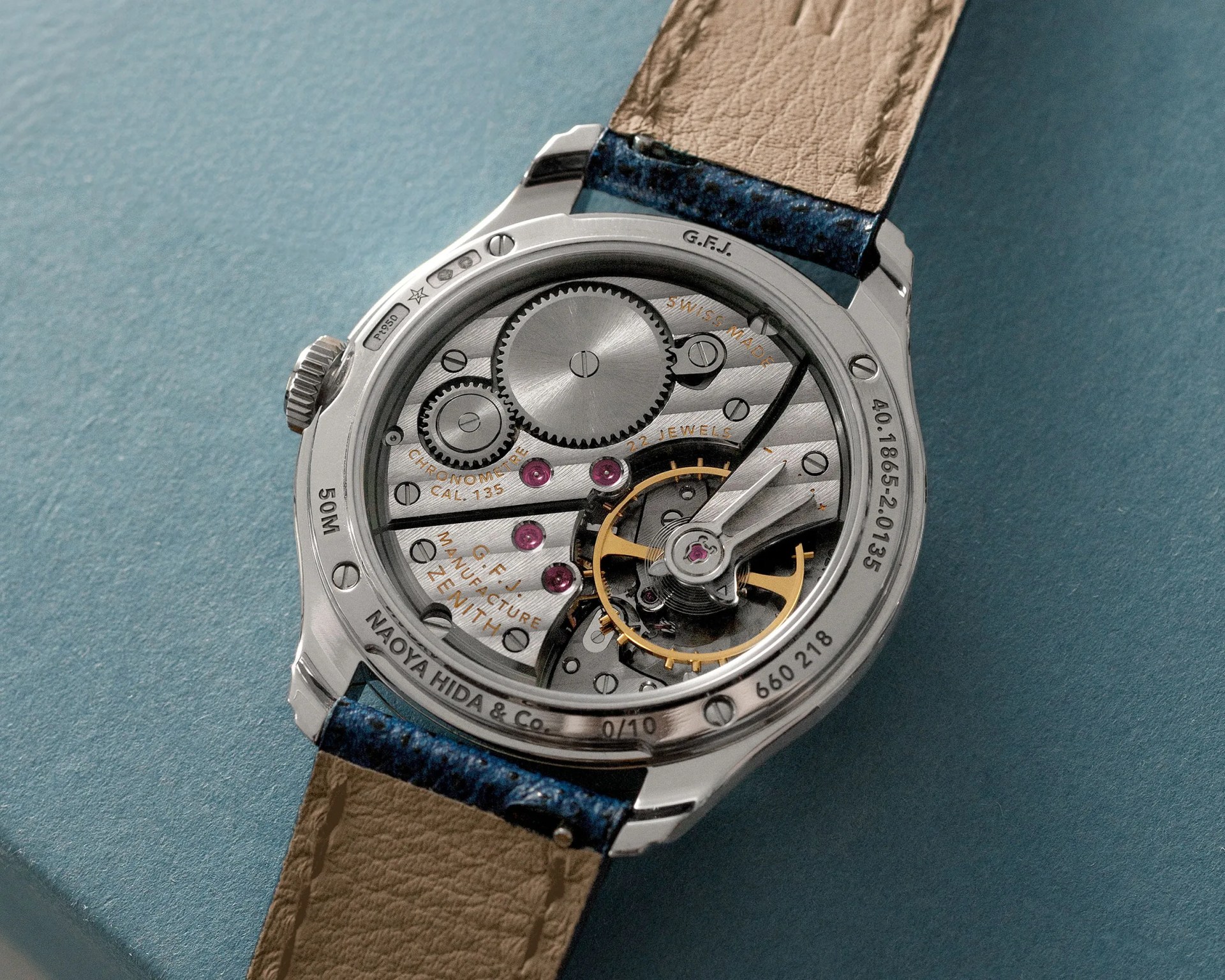Close-up of the back of a mechanical wristwatch showing gears and inscriptions, with a beige leather strap and blue stitching.