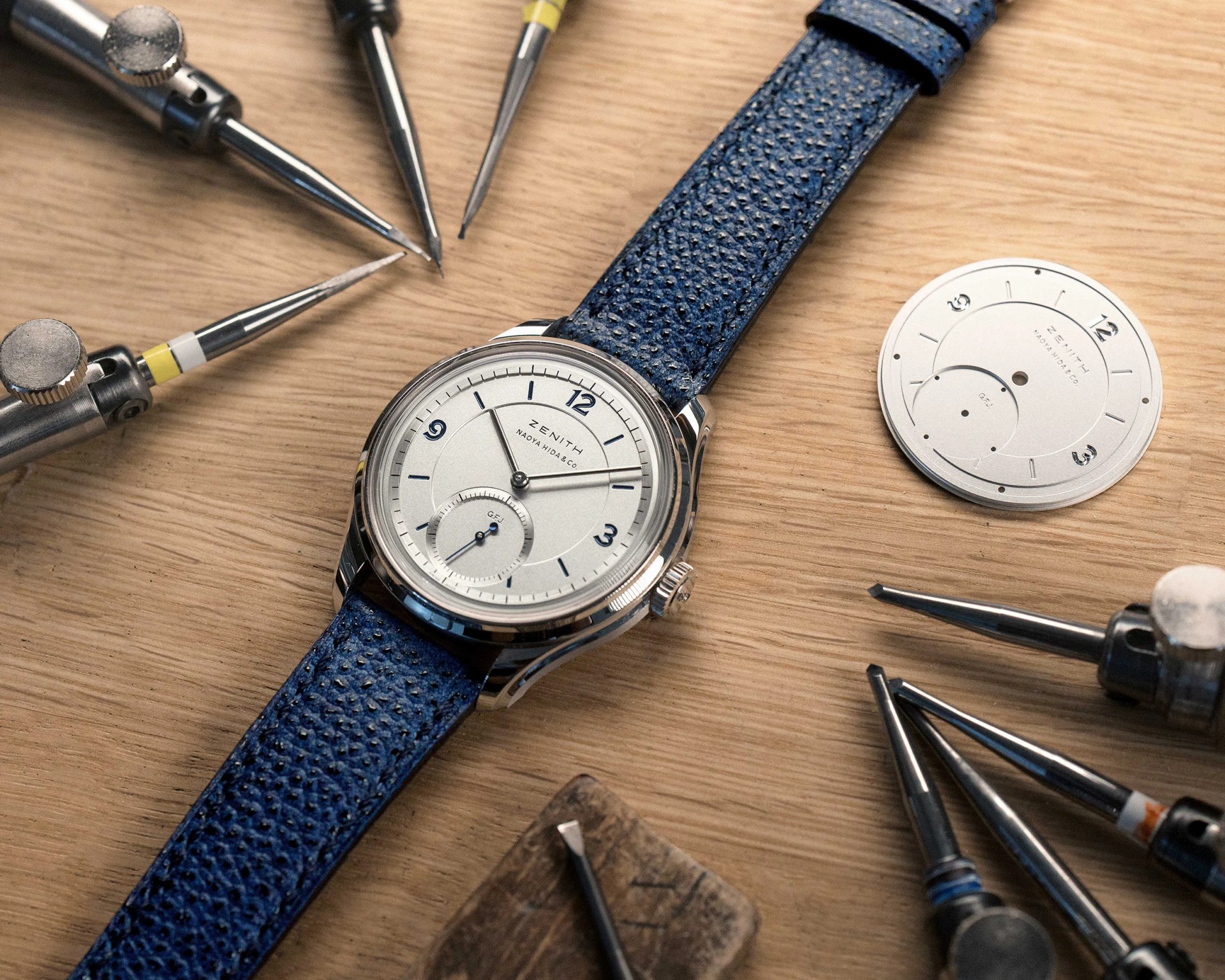 Silver Zenith wristwatch with white dial and blue leather strap on a wooden surface surrounded by watchmaking tools.