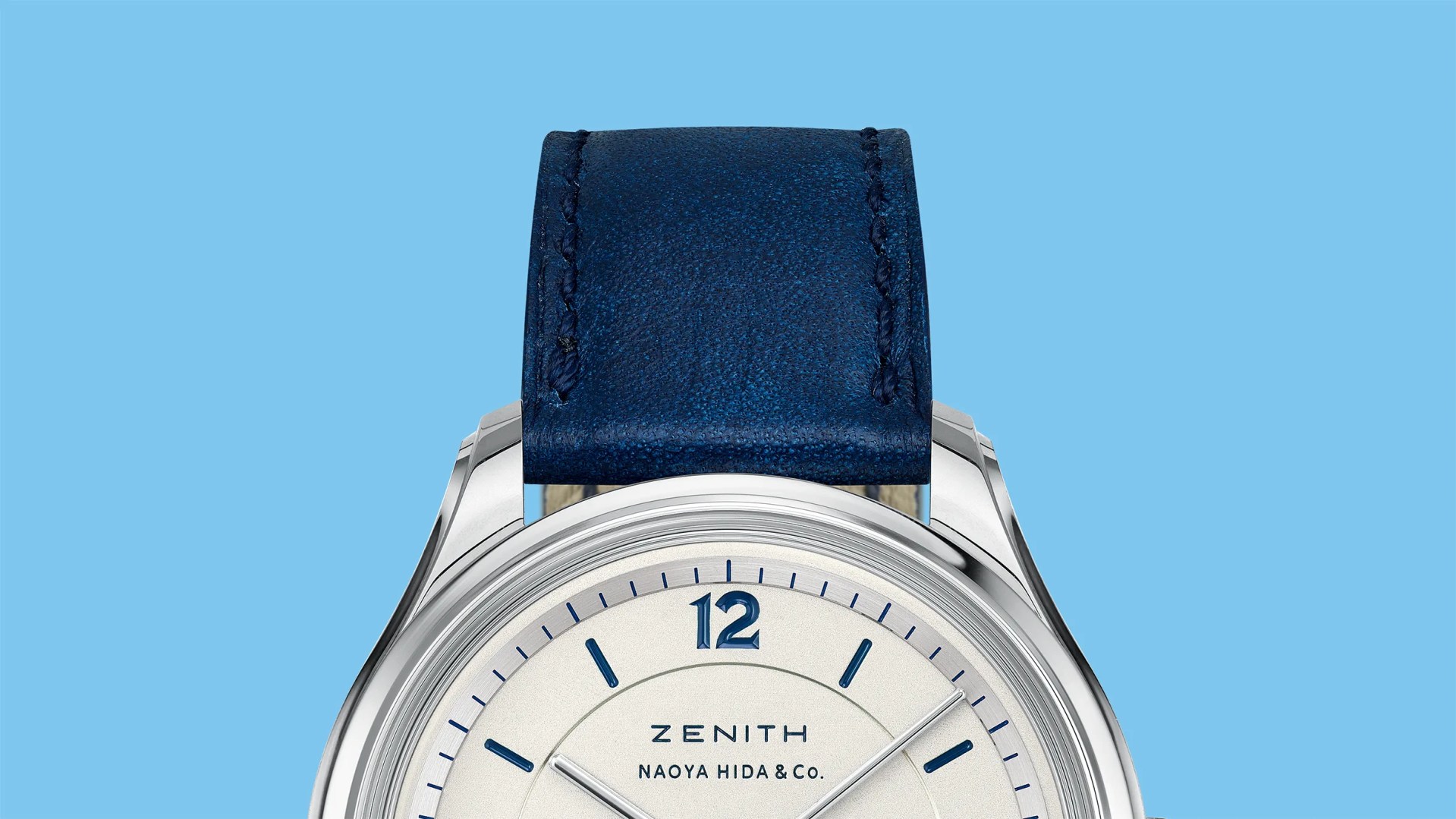 Close-up of a silver Zenith watch with a white dial, blue hour markers, and a blue leather strap against a blue background.