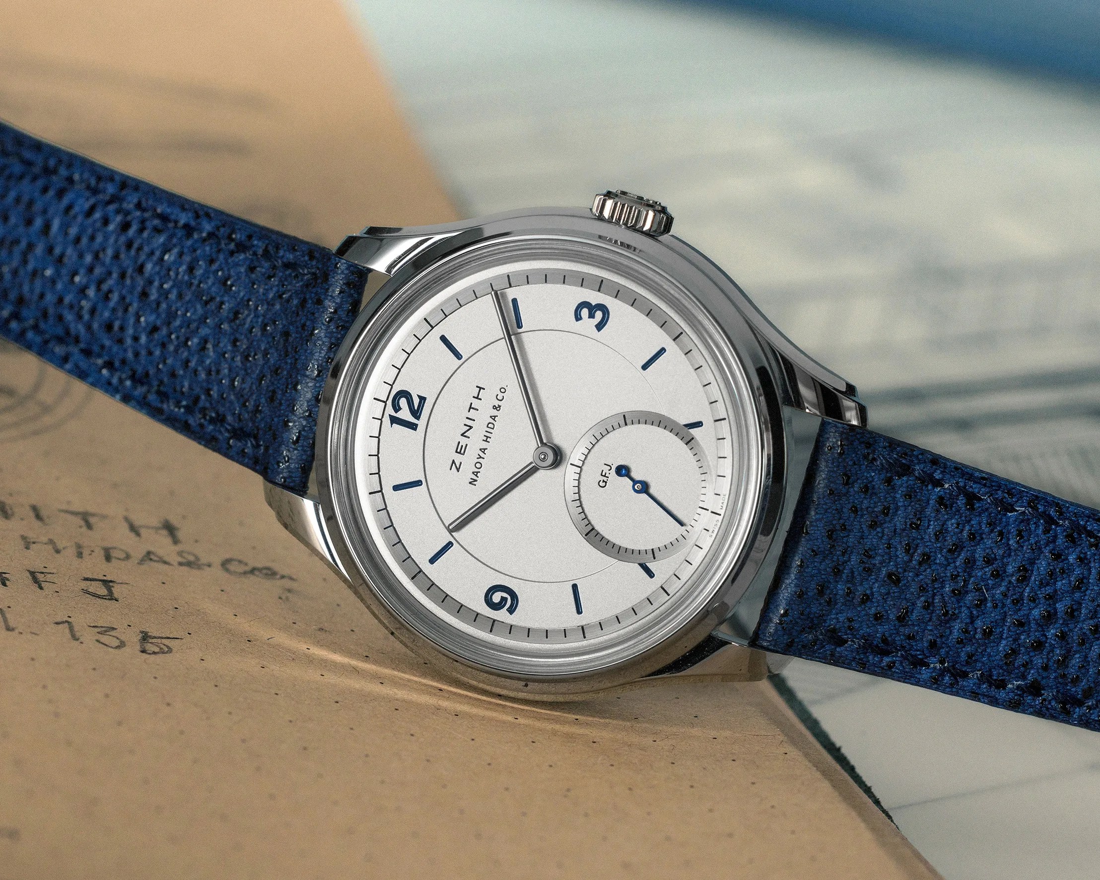 Silver Zenith wristwatch with white dial, blue numerals, and blue textured leather strap on a book.