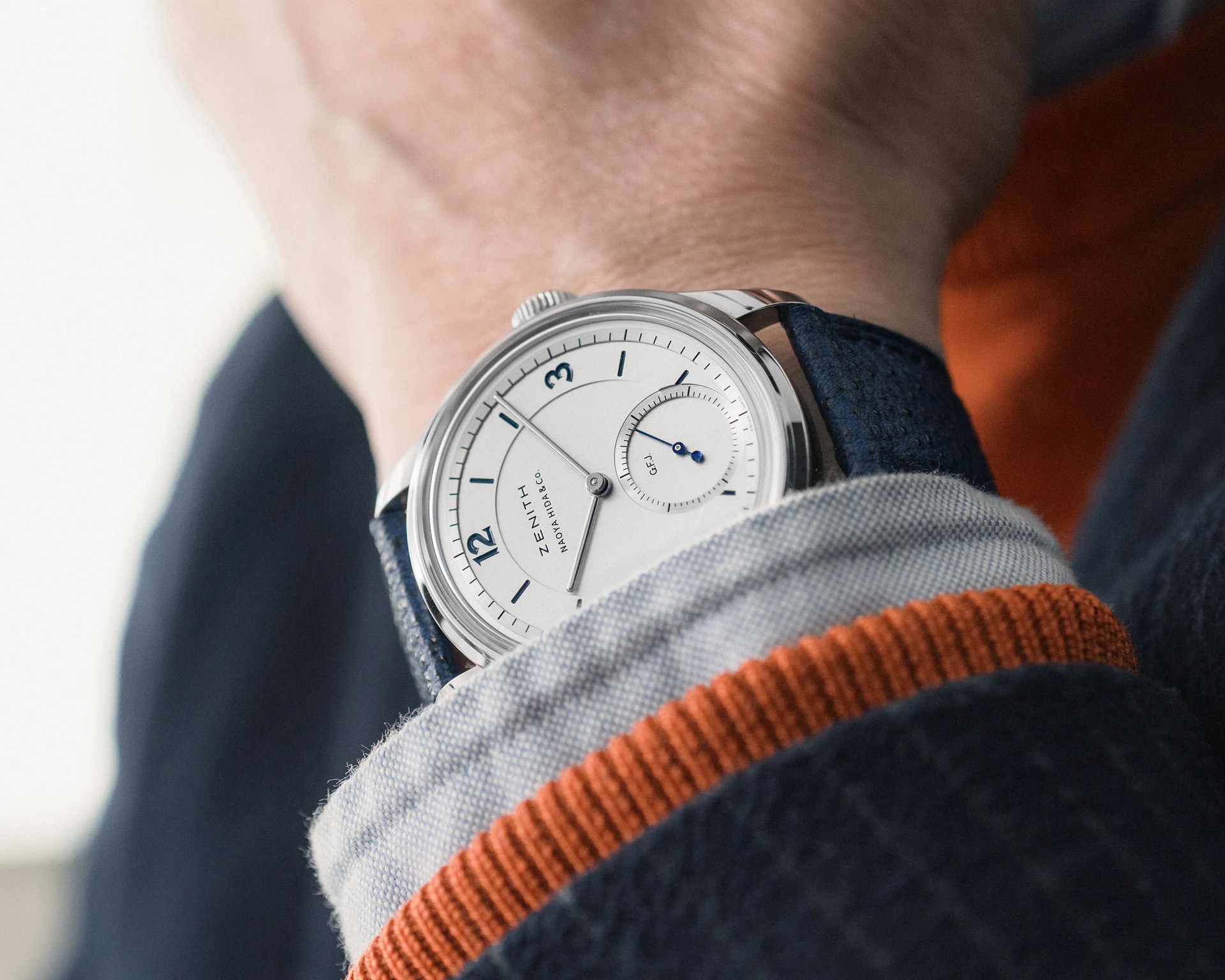 Silver Zenith wristwatch with white dial and blue leather strap worn under a shirt cuff with orange trim.