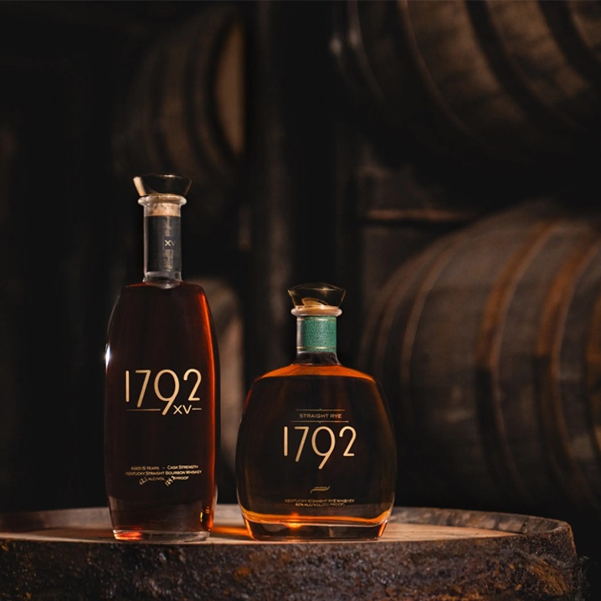 Two bottles of 1792 bourbon whiskey on a wooden barrel with stacked barrels in the background.