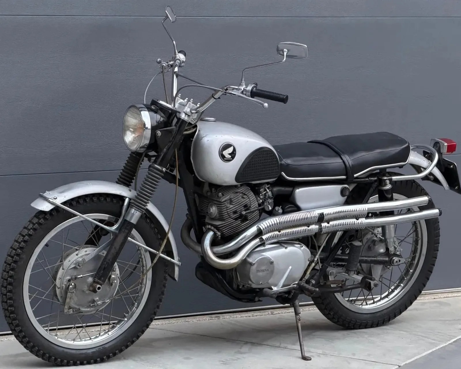 Silver and black vintage Honda motorcycle with knobby tires and dual exhaust pipes parked on a concrete floor.