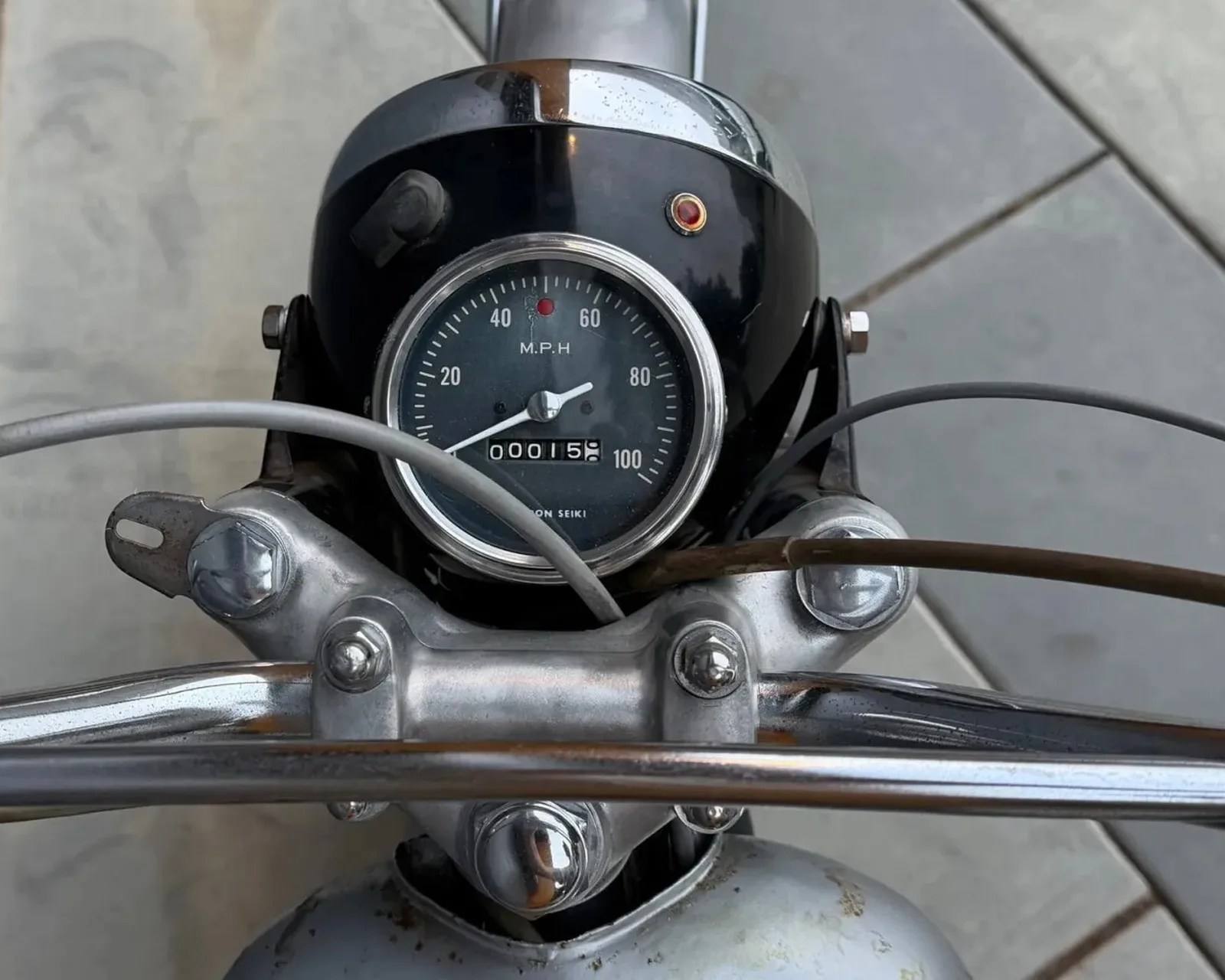 Close-up of a vintage motorcycle speedometer showing 15 miles and chrome handlebars with cables.