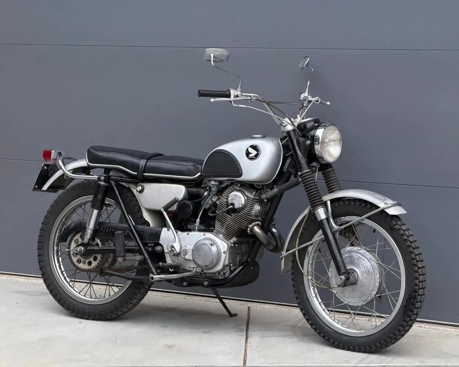 Vintage silver and black Honda motorcycle with knobby tires parked on concrete against a gray wall.