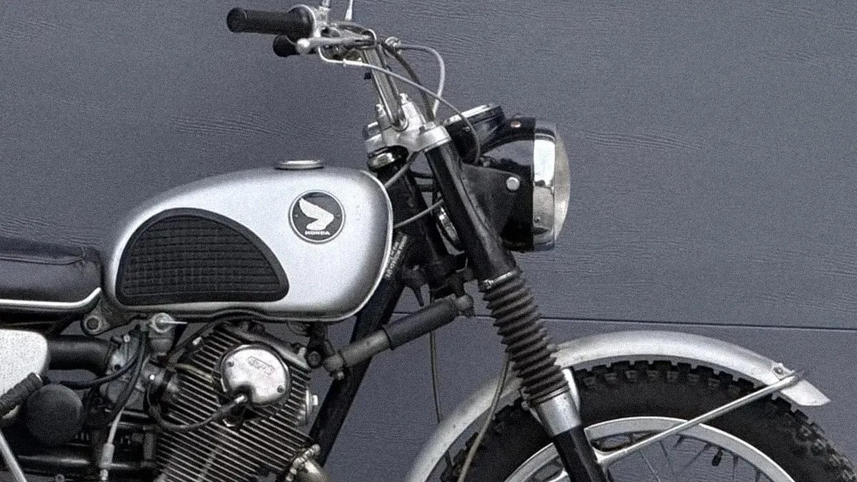 Silver and black vintage Honda motorcycle with a round headlight and knobby tires against a gray wall.