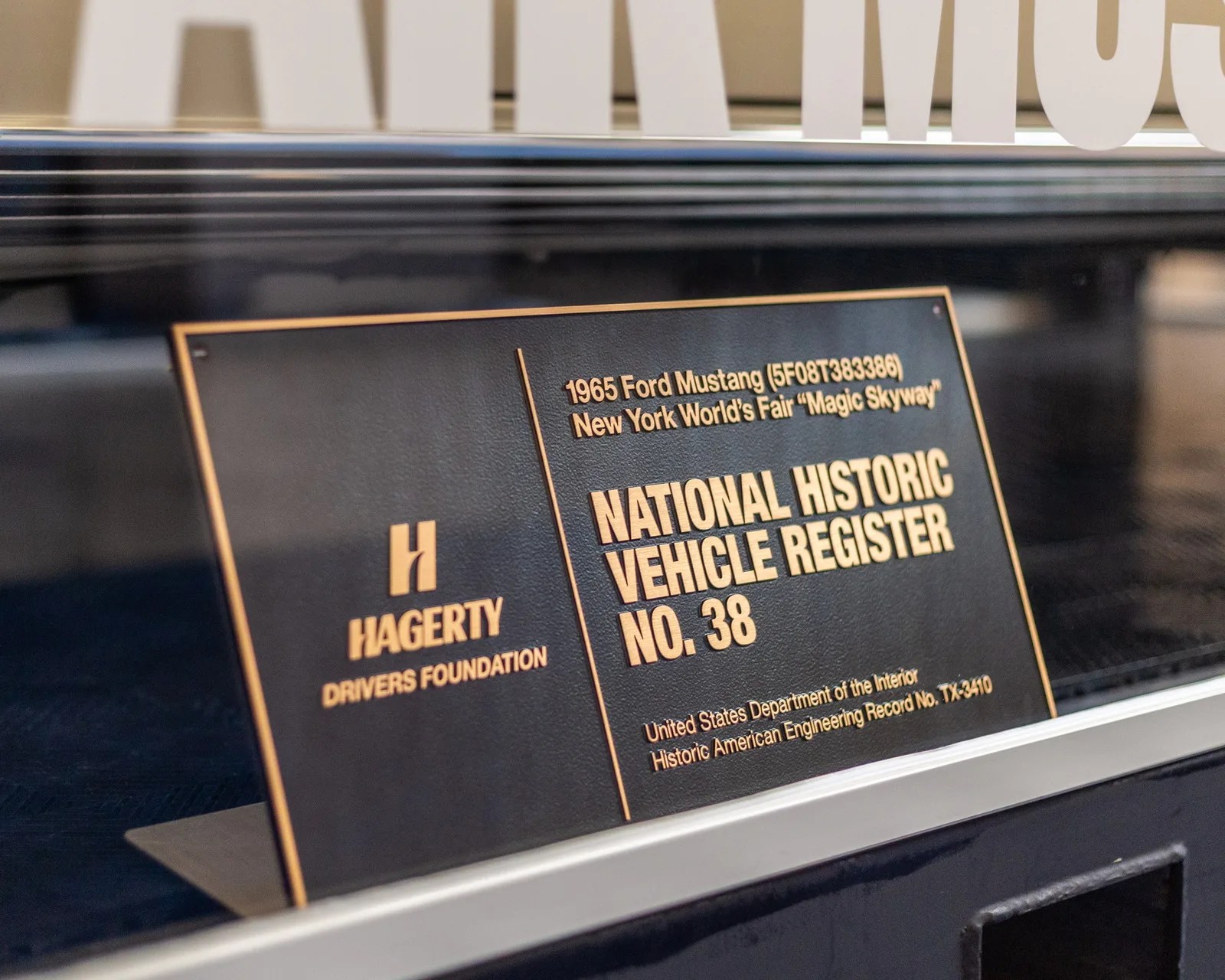 Black and gold plaque reading "1965 Ford Mustang (5F08T383386) New York World's Fair 'Magic Skyway' National Historic Vehicle Register No. 38" on a display.