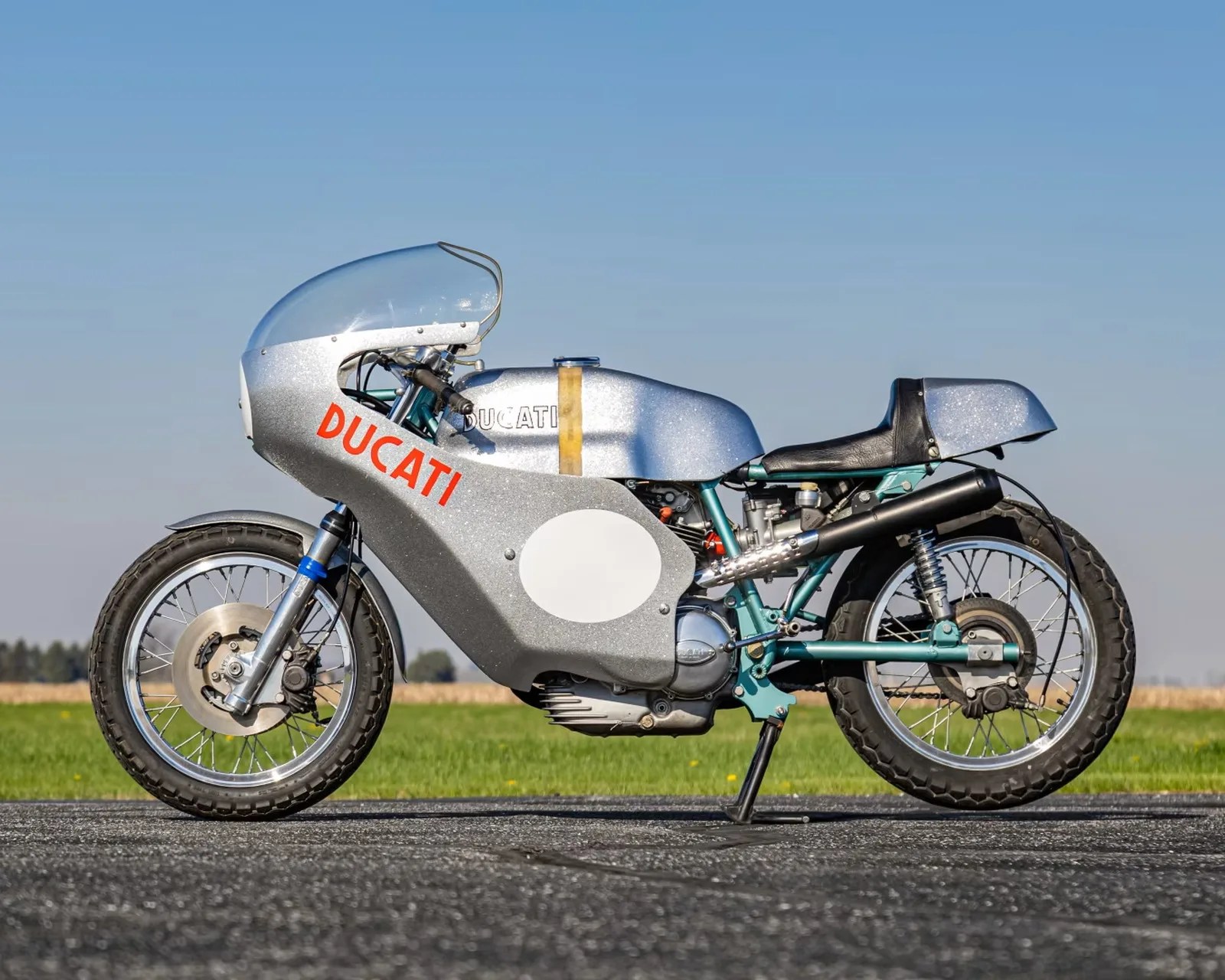Silver vintage Ducati racing motorcycle with red logo and clear windshield on a paved road.