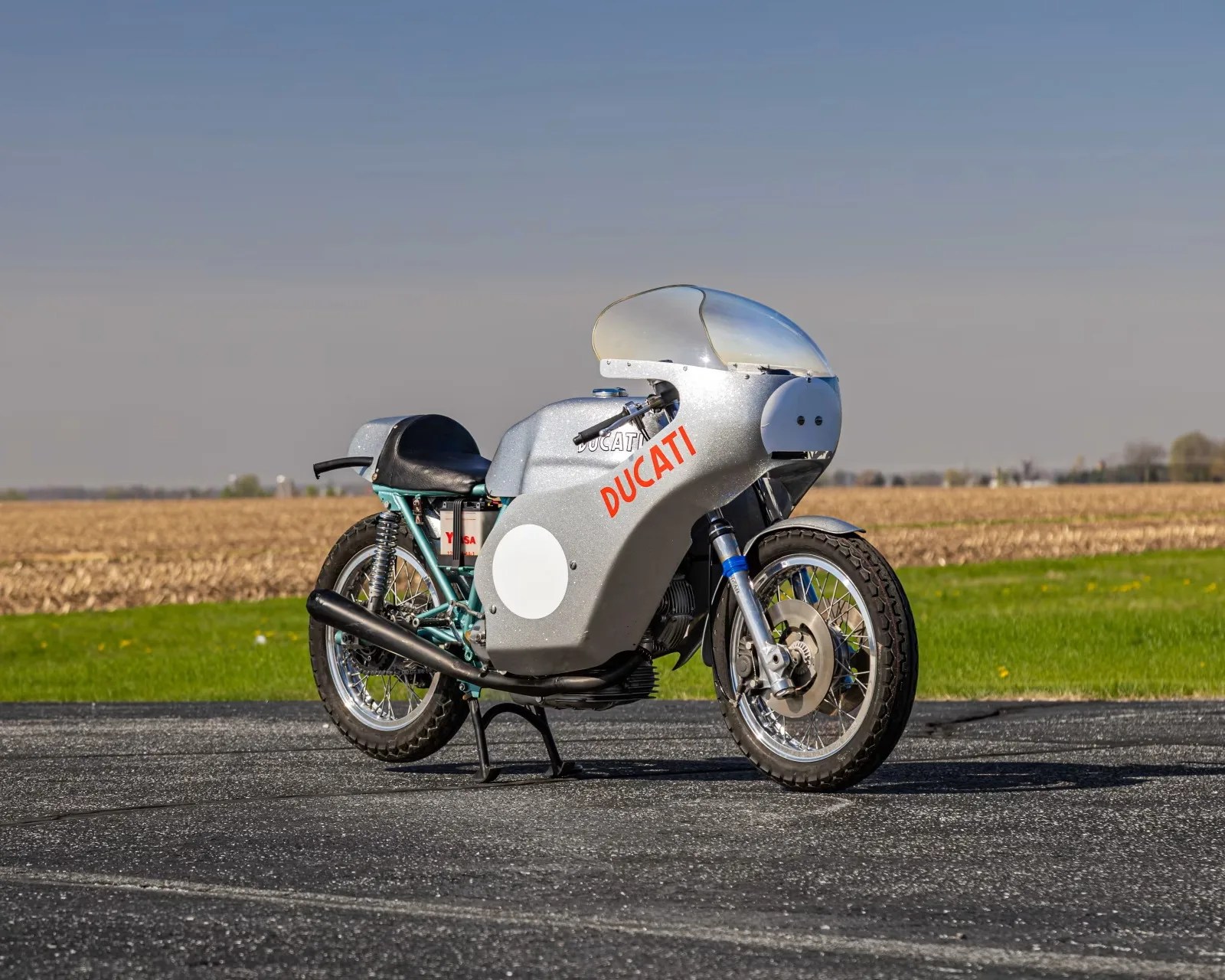 Silver vintage Ducati racing motorcycle with black seat and clear windshield on asphalt with field background.
