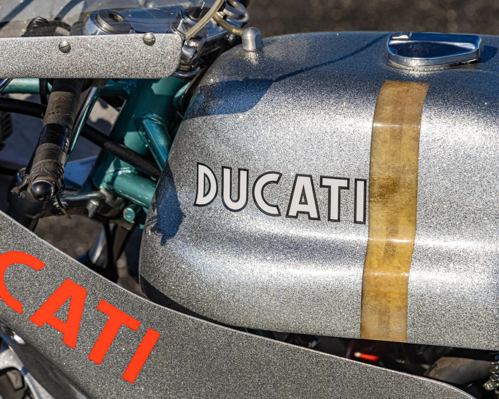 Silver glitter-finish Ducati motorcycle fuel tank with a vertical brown tape strip and part of the handlebar visible.