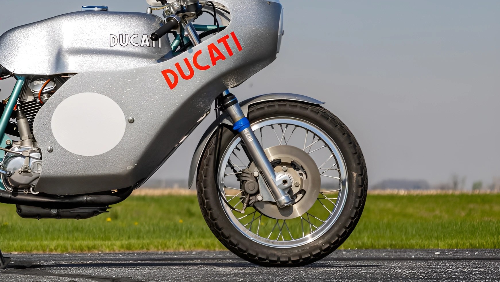 Silver Ducati motorcycle front wheel and part of the body with red Ducati logo on a road with grass background.