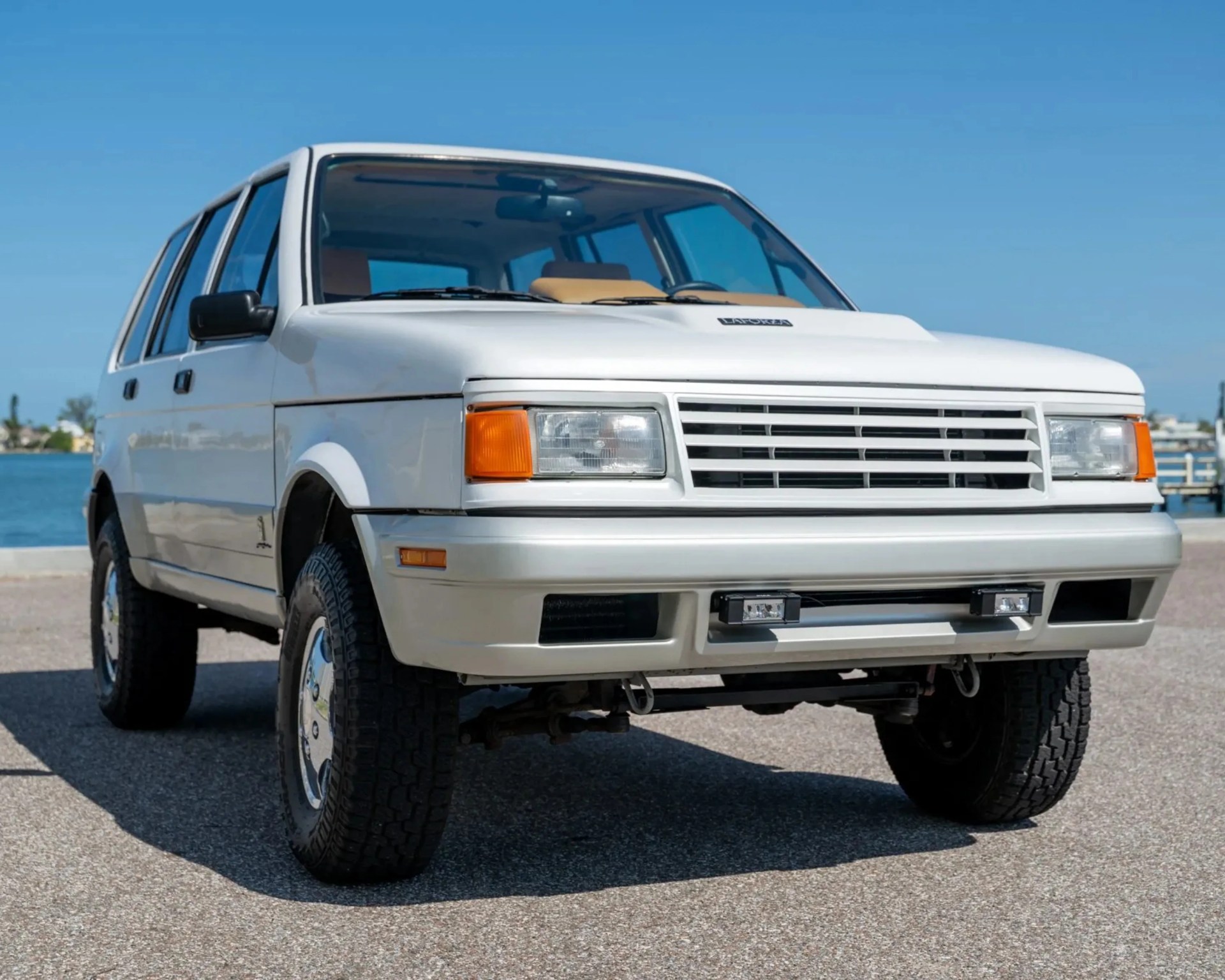 White vintage SUV with rectangular headlights and a black side mirror parked on pavement near water.