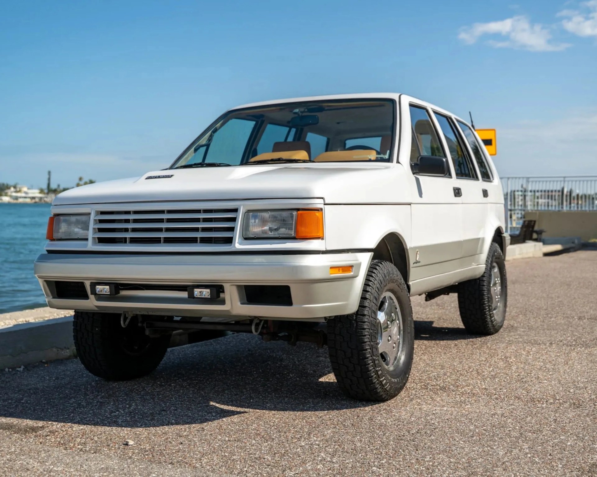 White vintage SUV with tan interior parked on a waterfront road under a clear blue sky.