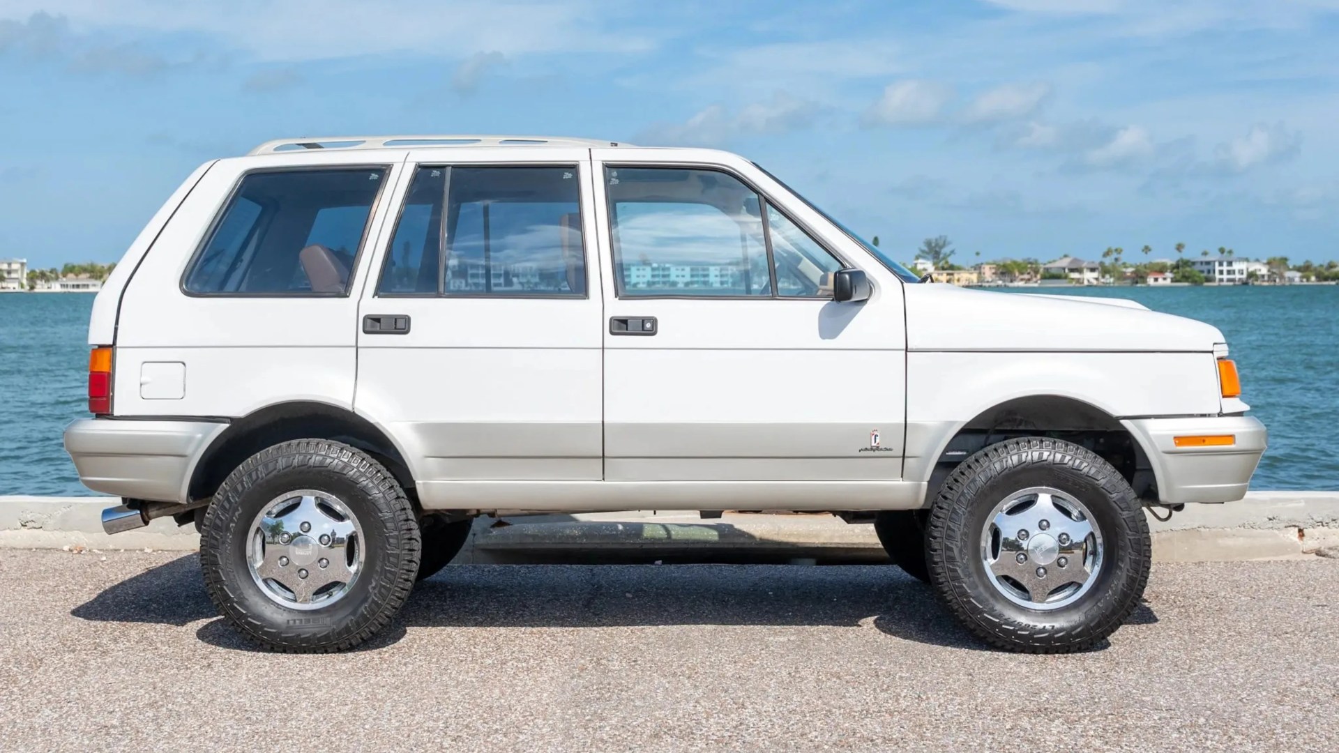 White and silver 1990s SUV with chrome wheels parked by a waterfront under a blue sky.