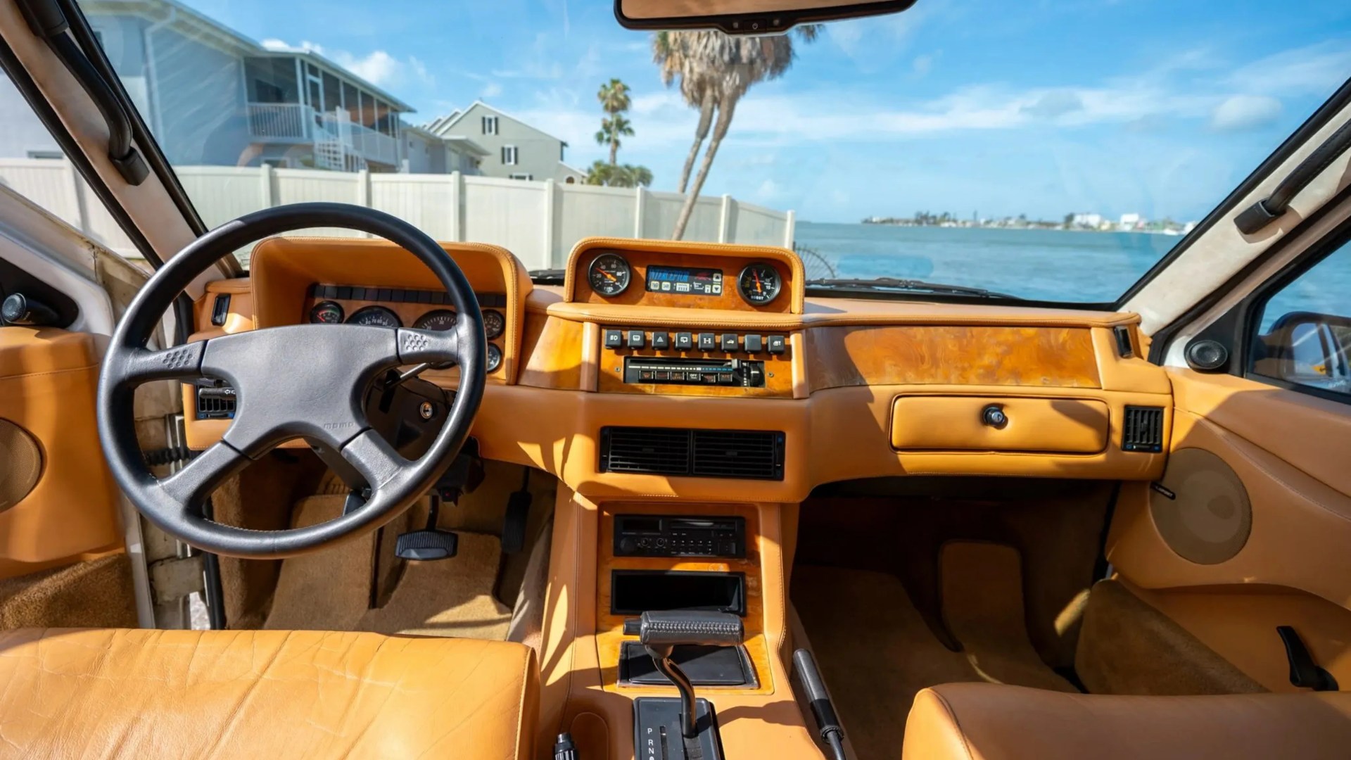 Vintage car interior with tan leather seats, wood grain dashboard, and black steering wheel facing a waterfront view.