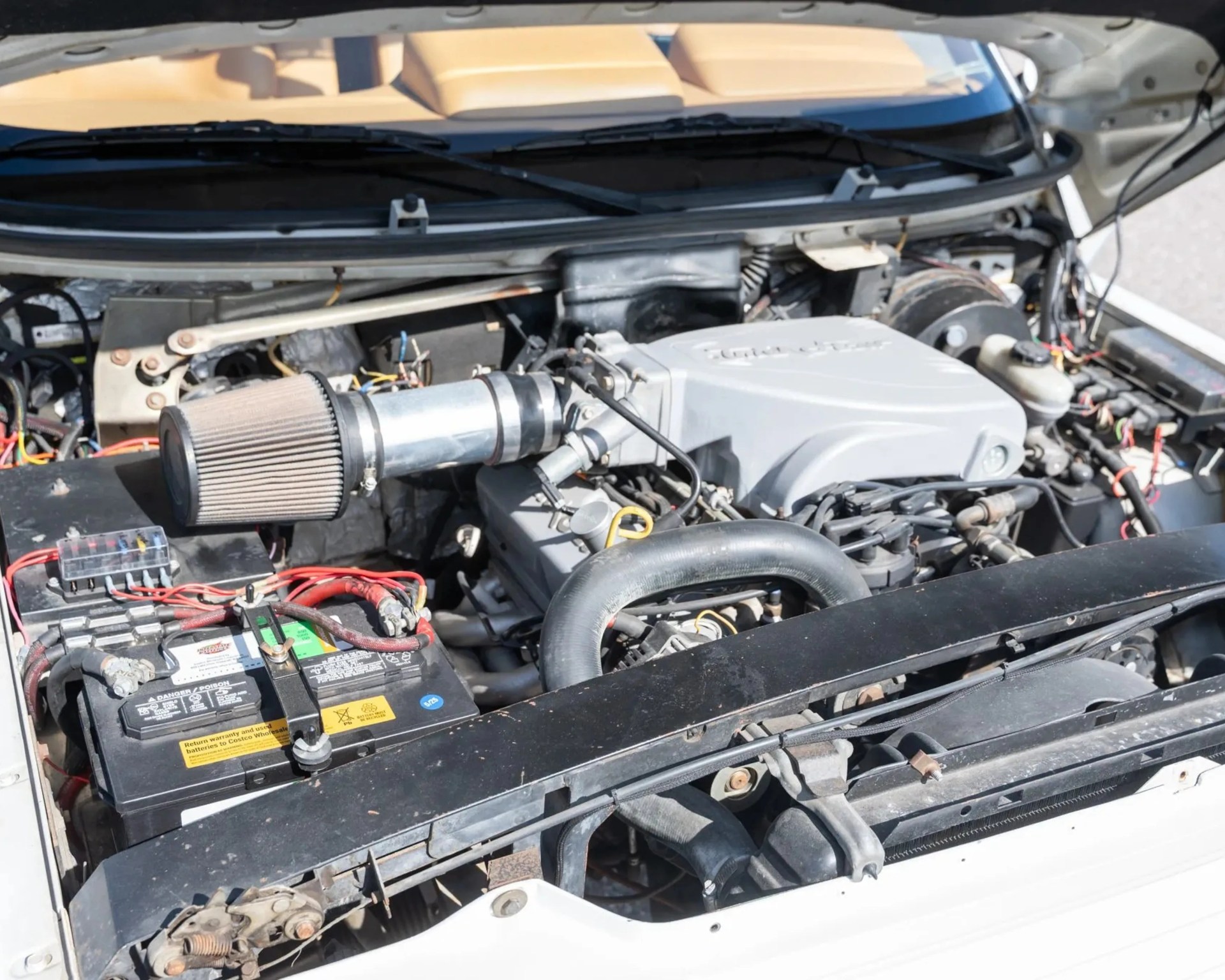 Car engine bay with a visible air filter, battery, and various engine components under an open hood.