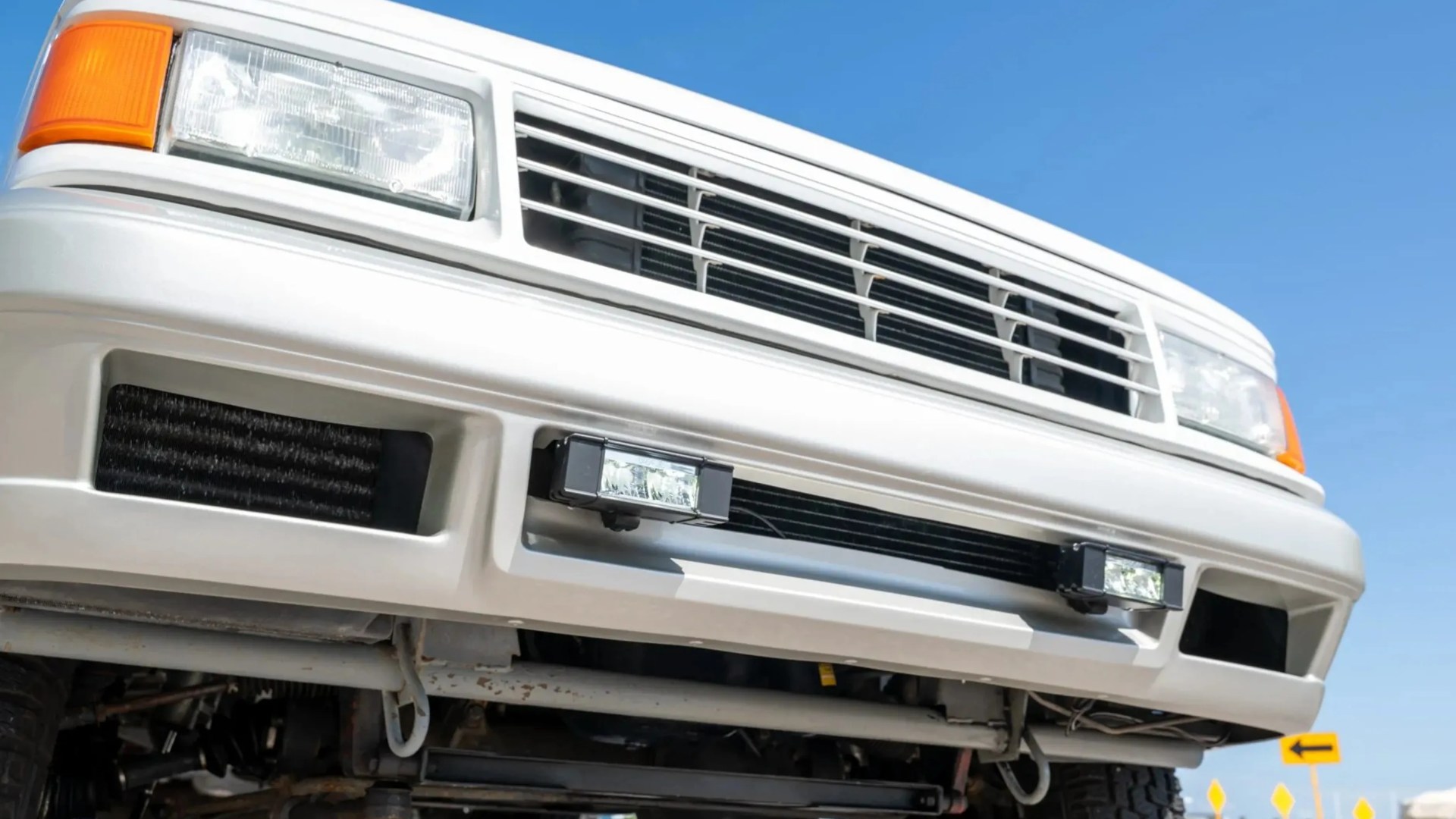 Close-up of a white vehicle's front bumper with rectangular headlights, grille, and two small rectangular fog lights.