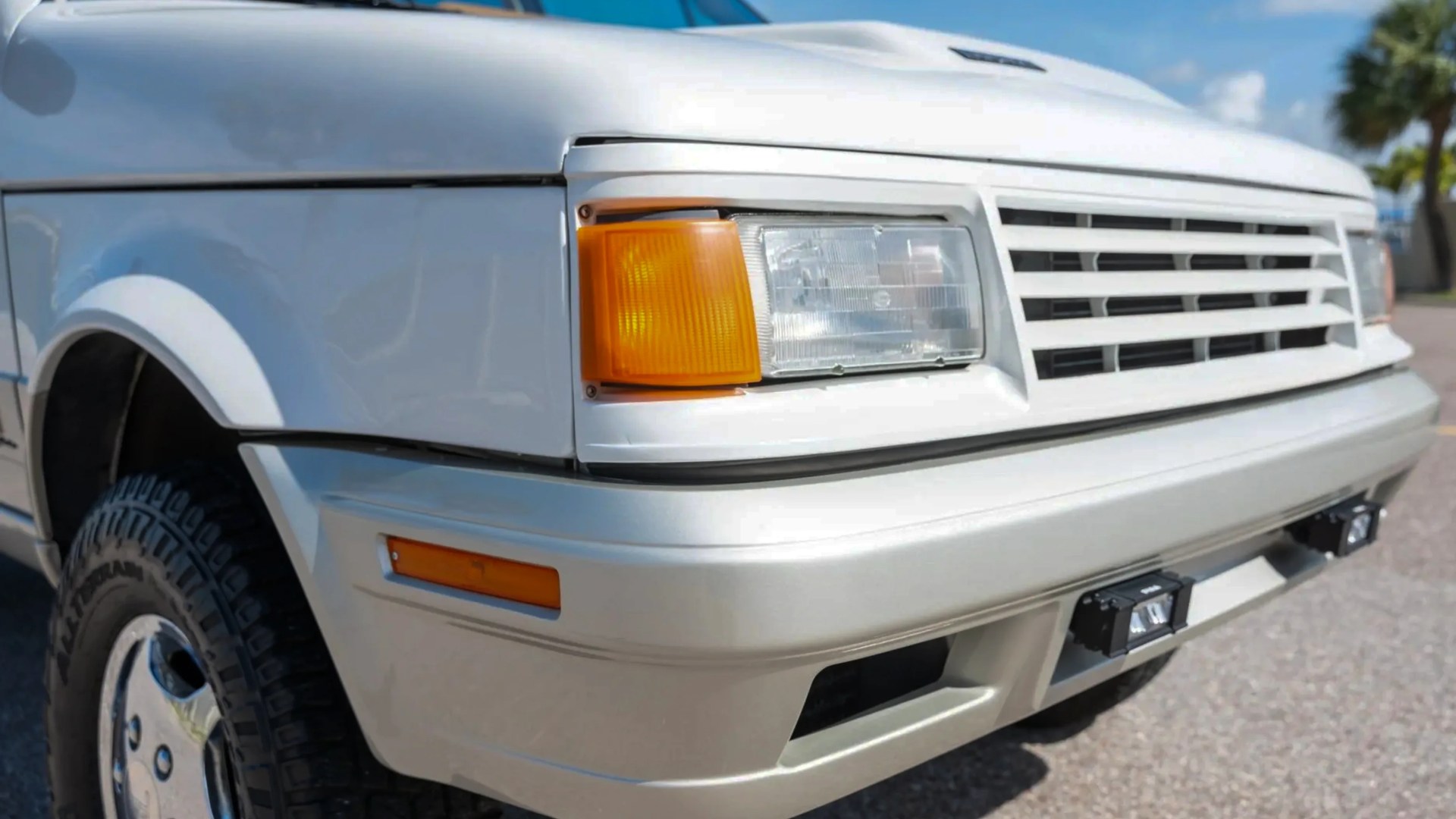 Close-up of the front left corner of a white vehicle showing the headlight, amber turn signal, grille, and tire.