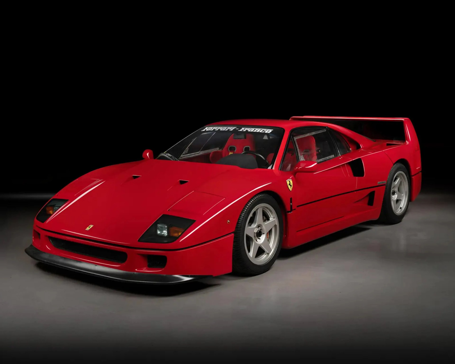 Red Ferrari F40 sports car with a large rear wing and silver five-spoke wheels on a gray floor.