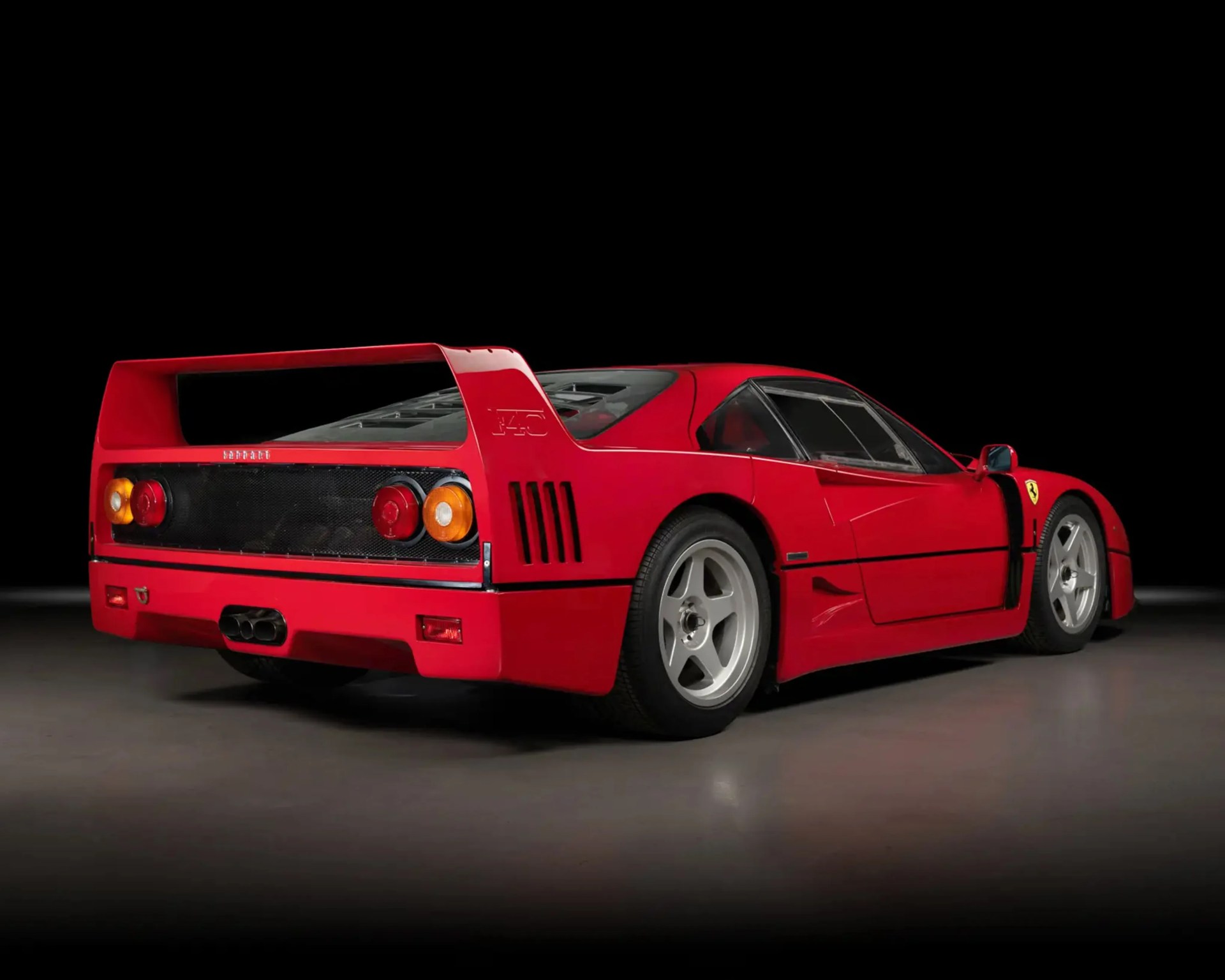 Red Ferrari F40 sports car rear with large rear wing and round taillights on gray floor and black background.