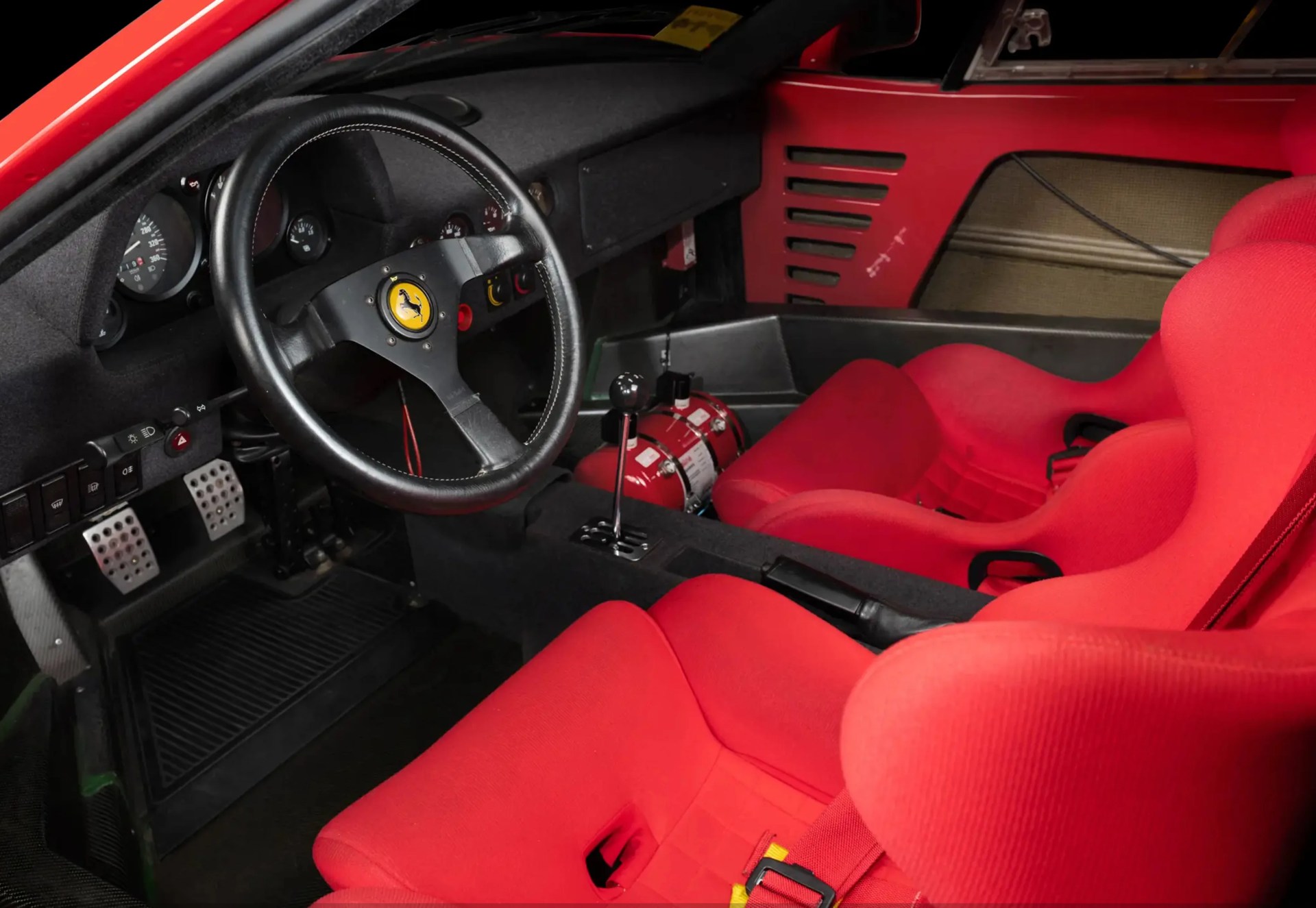 Black leather steering wheel with yellow Ferrari logo, red racing bucket seats, and manual gear shifter inside a red sports car.