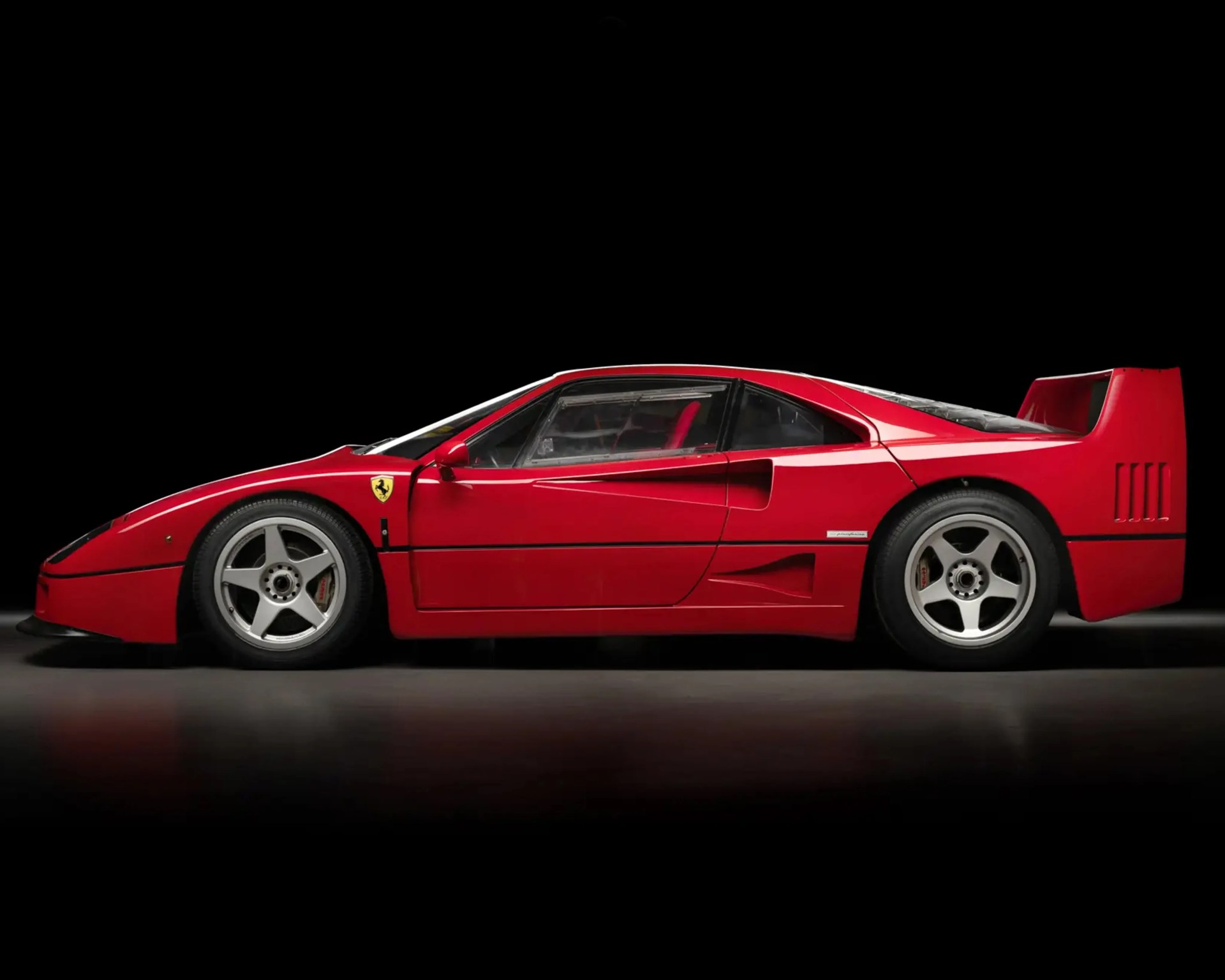 Red Ferrari F40 sports car side profile with Pininfarina badge and five-spoke silver wheels on black background.