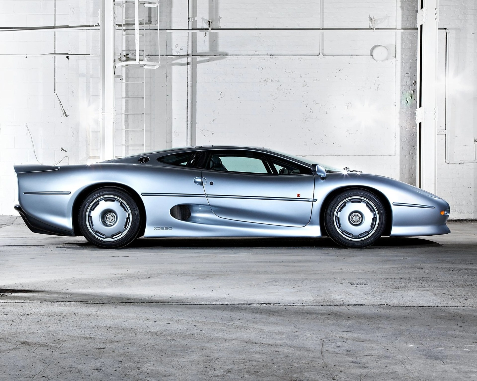 Silver Jaguar XJ220 sports car parked indoors against a white brick wall.