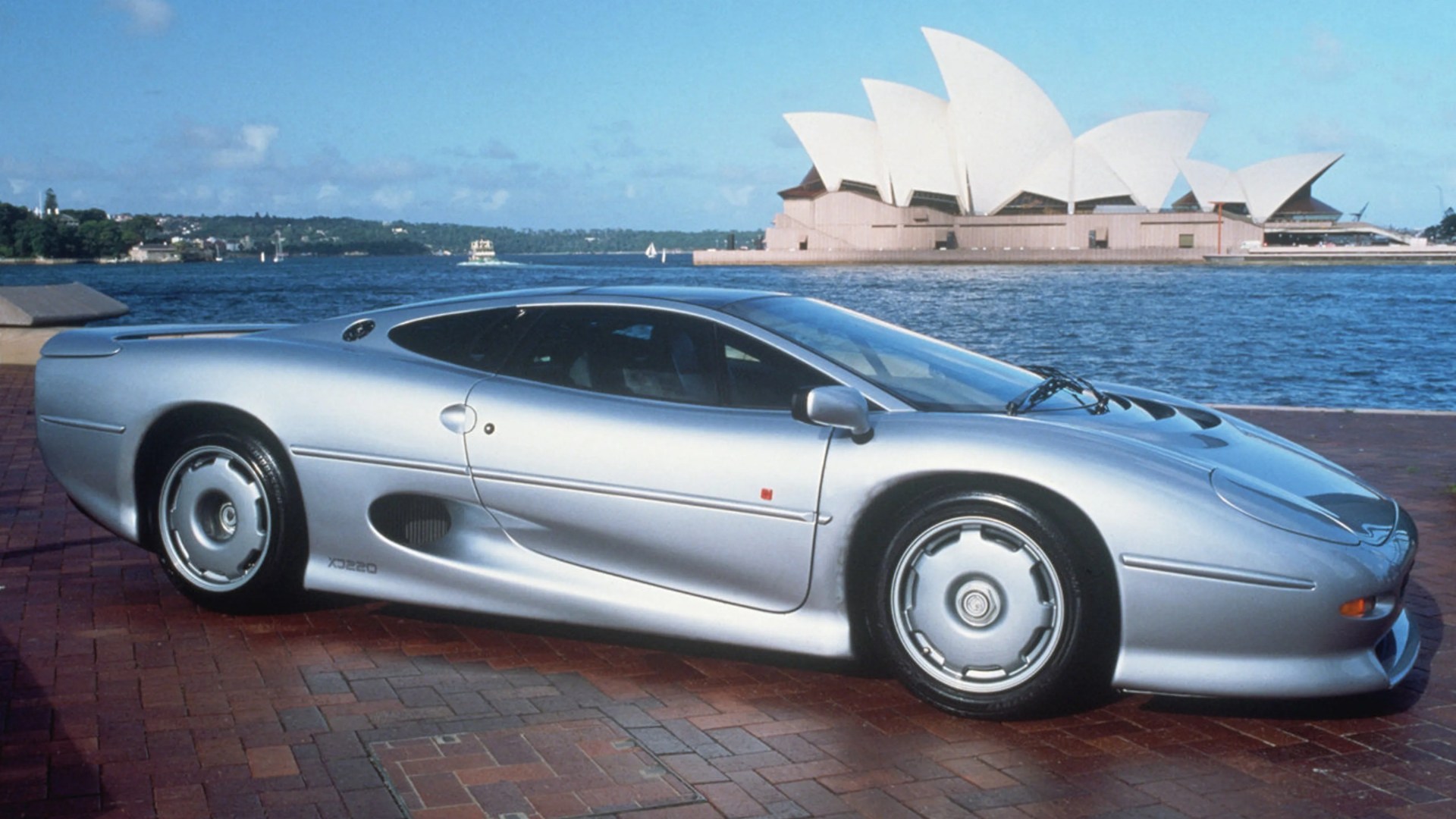 Silver Jaguar XJ220 sports car parked on brick pavement with Sydney Opera House and water in the background.
