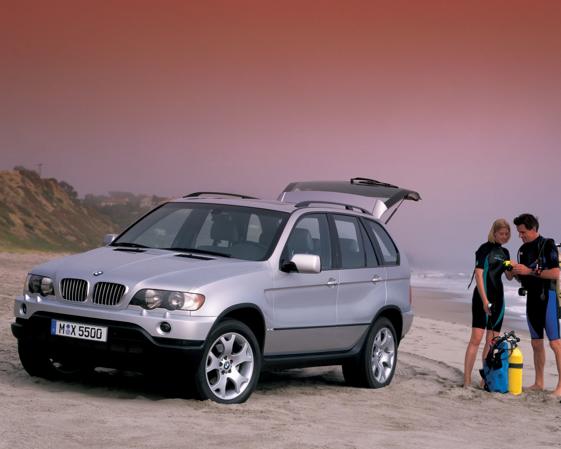 Silver BMW X5 SUV parked on a sandy beach with two people in wetsuits preparing scuba gear nearby.
