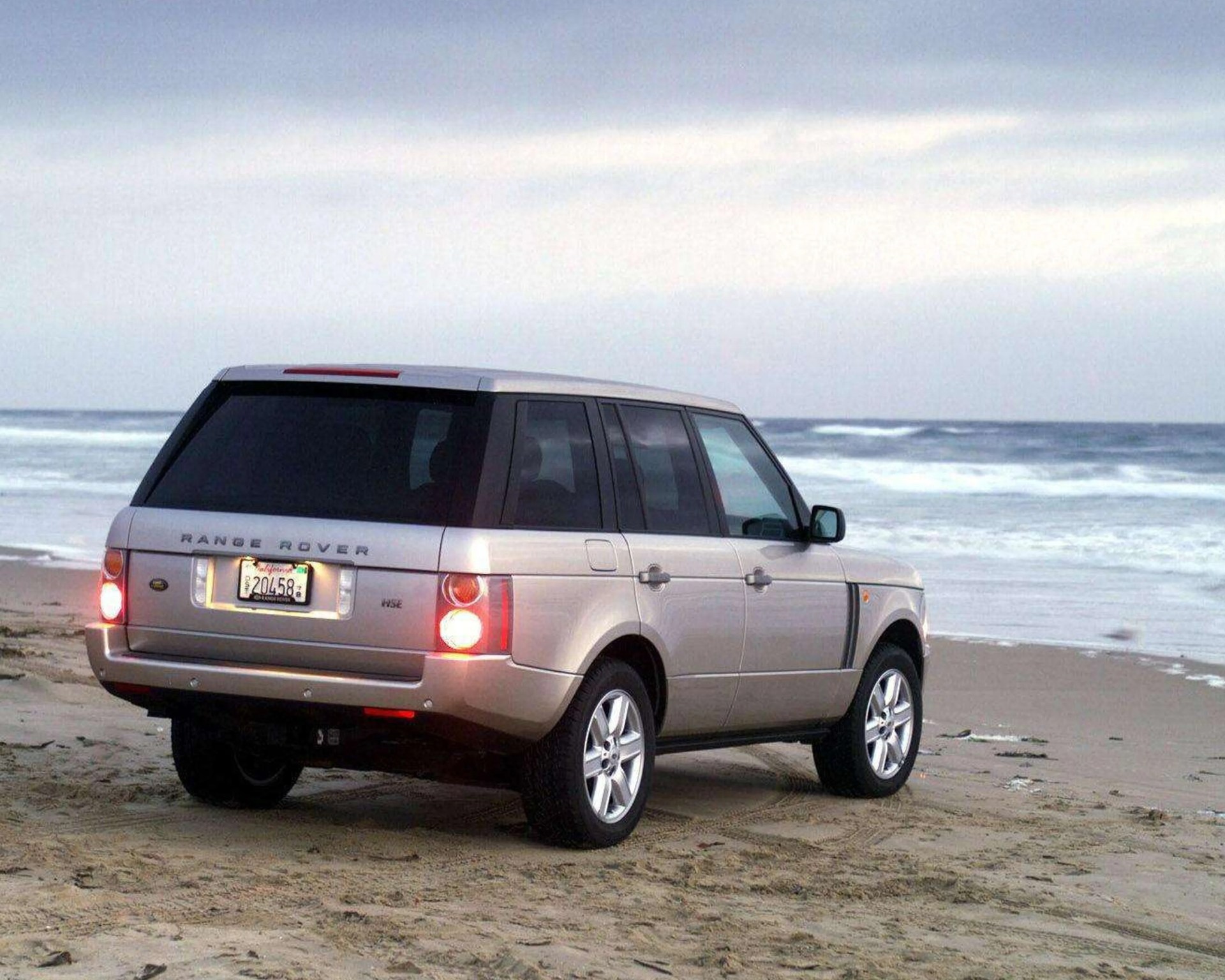 Silver Range Rover HSE SUV parked on sandy beach near ocean waves under cloudy sky.